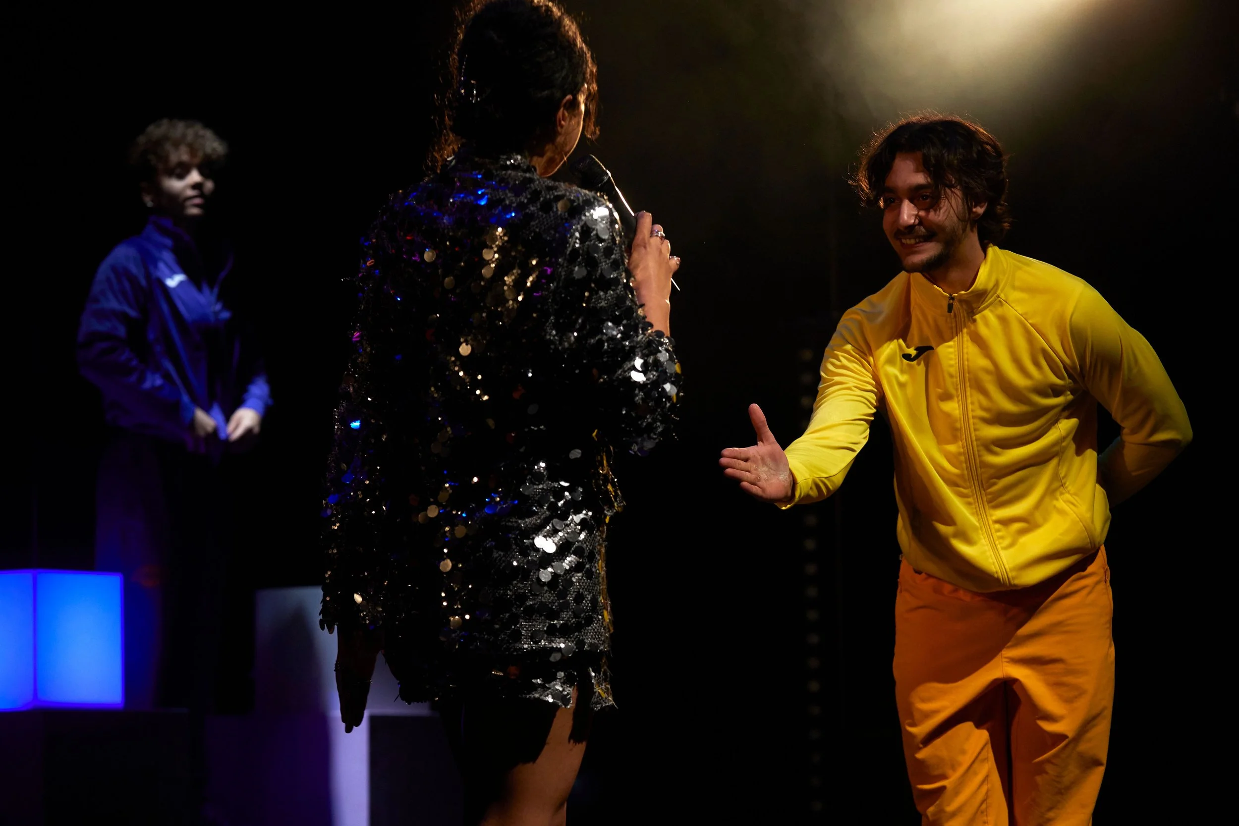 Three people on stage, one woman in a sequined blazer talking into a microphone, a man in a yellow tracksuit reaching out to her, another woman in a blue tracksuit in the background.