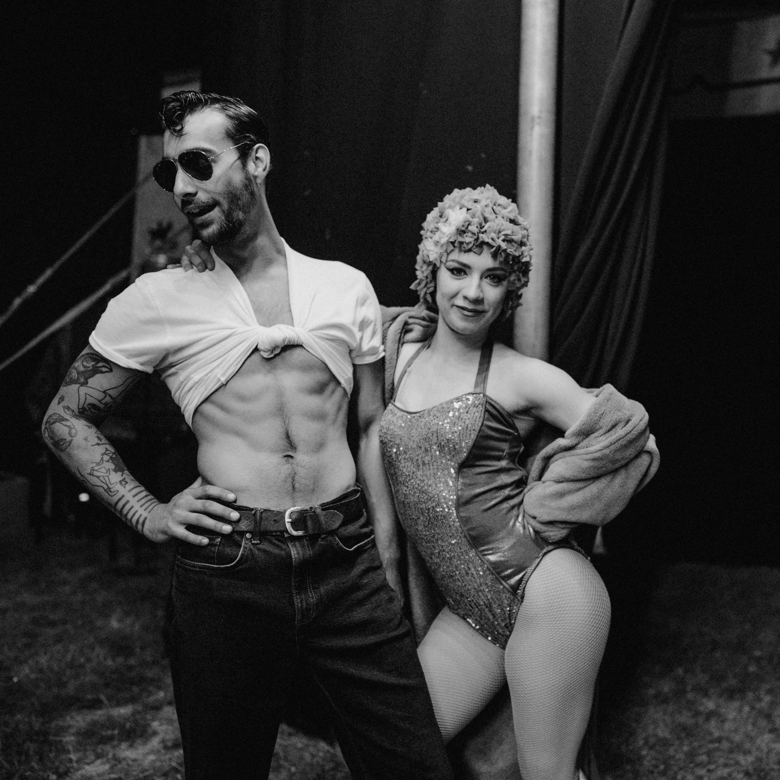 A man with sunglasses and tattoos, wearing a tied-up t-shirt revealing his chest and abs, standing next to a woman with a floral headpiece and a shiny costume, both posing backstage at a performance.