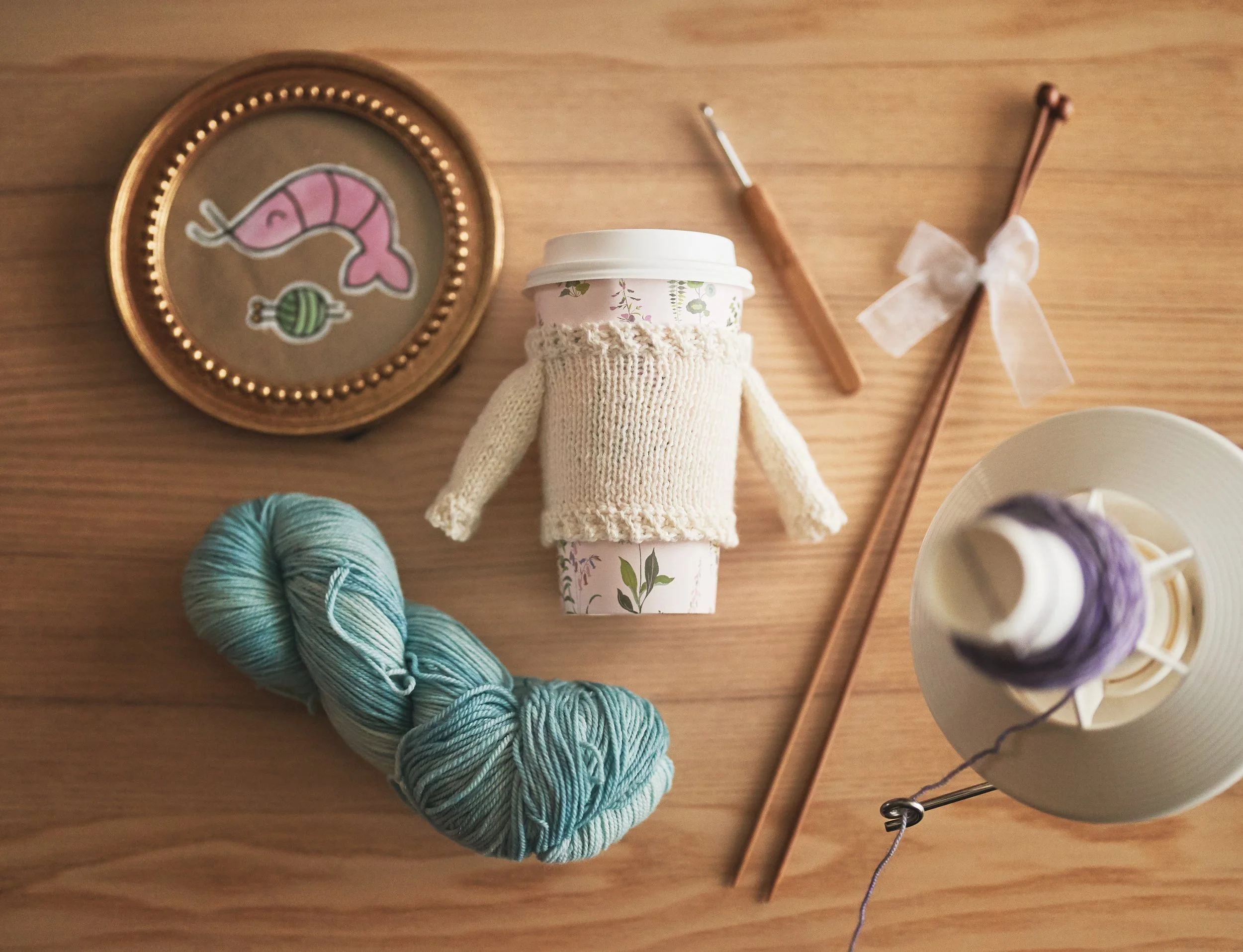 Flat lay of a disposable coffee cup with floral design, a knitted sleeve, and a paper cover, placed on a wooden surface surrounded by yarn, embroidery hoops, and a ceramic bowl with purple yarn.
