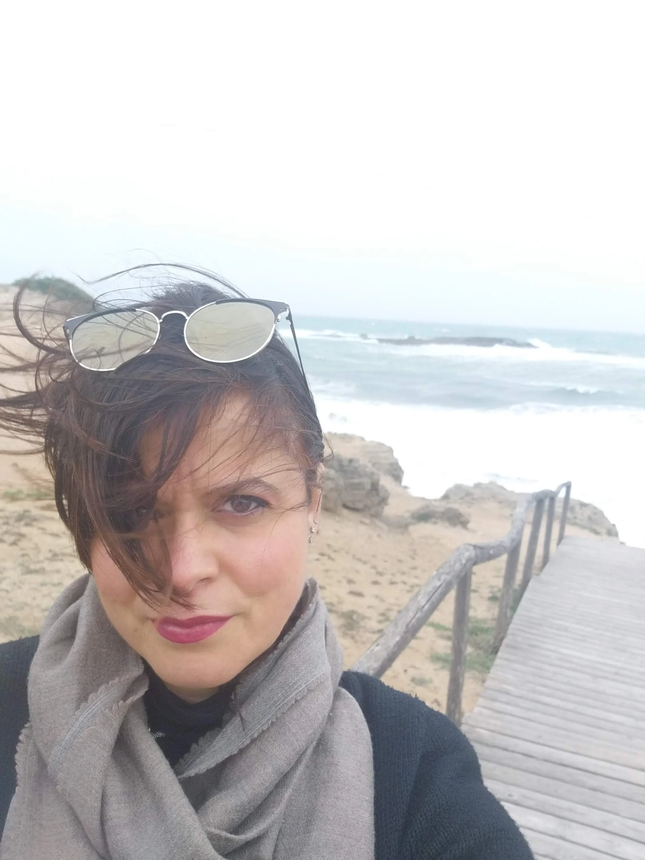 Donna con occhiali da sole sulla testa e sciarpa davanti a un mare ventoso con onde e una passerella di legno sulla spiaggia.