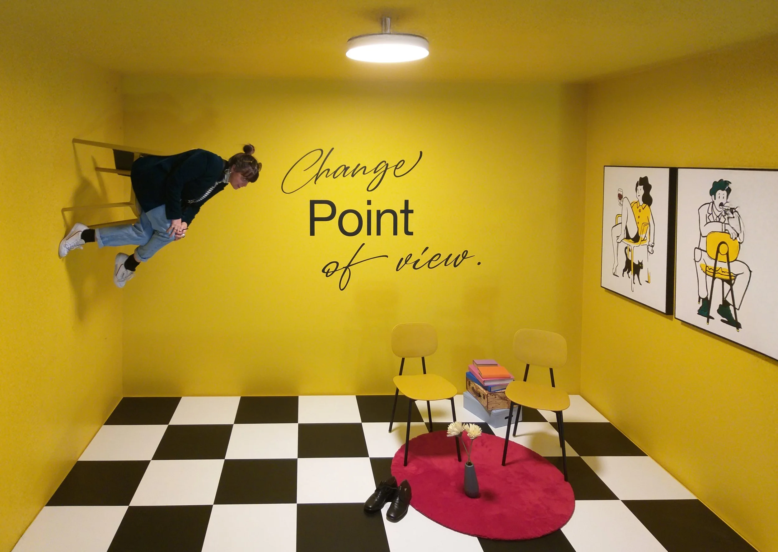 Stanza con pareti gialle, due sedie gialle con libro, tappeto rosso, vasi con fiori, foto di due donne sedute, scritta 'Change Point of view' sul muro.