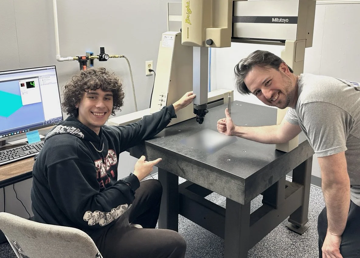 Kyle and student in a laboratory working with a large machine on a black table. One man with curly hair is sitting at a desk with a computer, and the other man with straight hair is standing, giving a thumbs up.