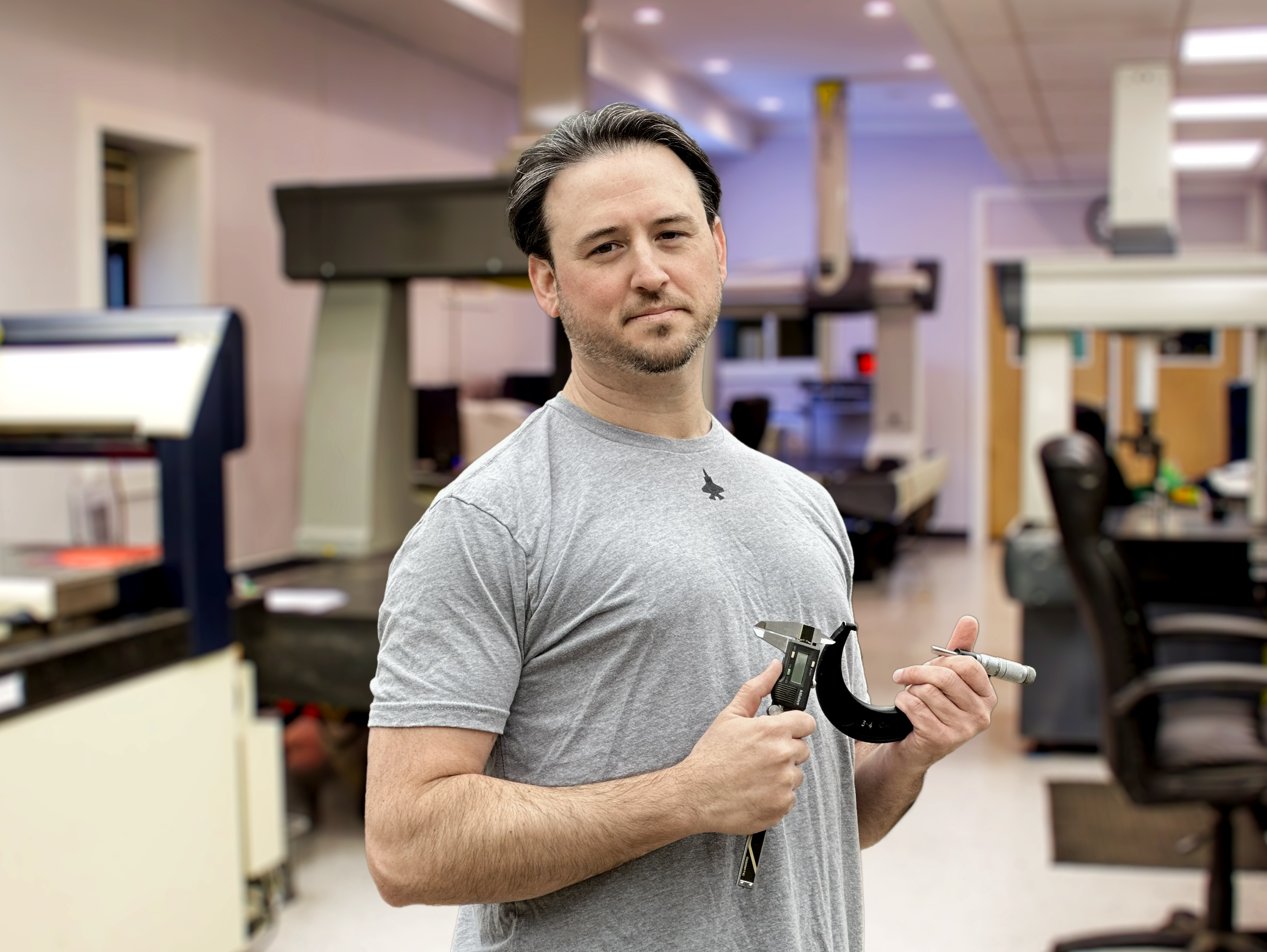 Kyle in gray t-shirt holding a caliper and a small screw inside a laboratory or workshop with scientific equipment and workbenches in the background.