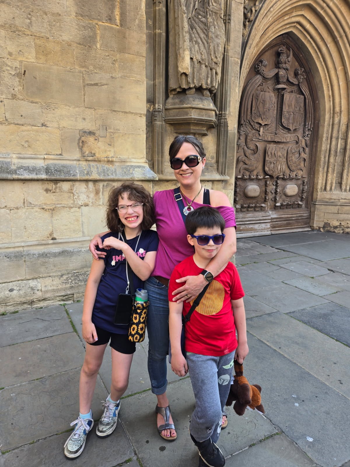 A woman and two children smiling and standing outside in front of a large ornate door on a historic stone building. The woman is wearing sunglasses, a purple top, and jeans. The girl has glasses, curly hair, a navy shirt with pink lettering, and shorts. The boy has sunglasses, a red shirt with a yellow circle, and gray sweatpants. They are posing happily together.