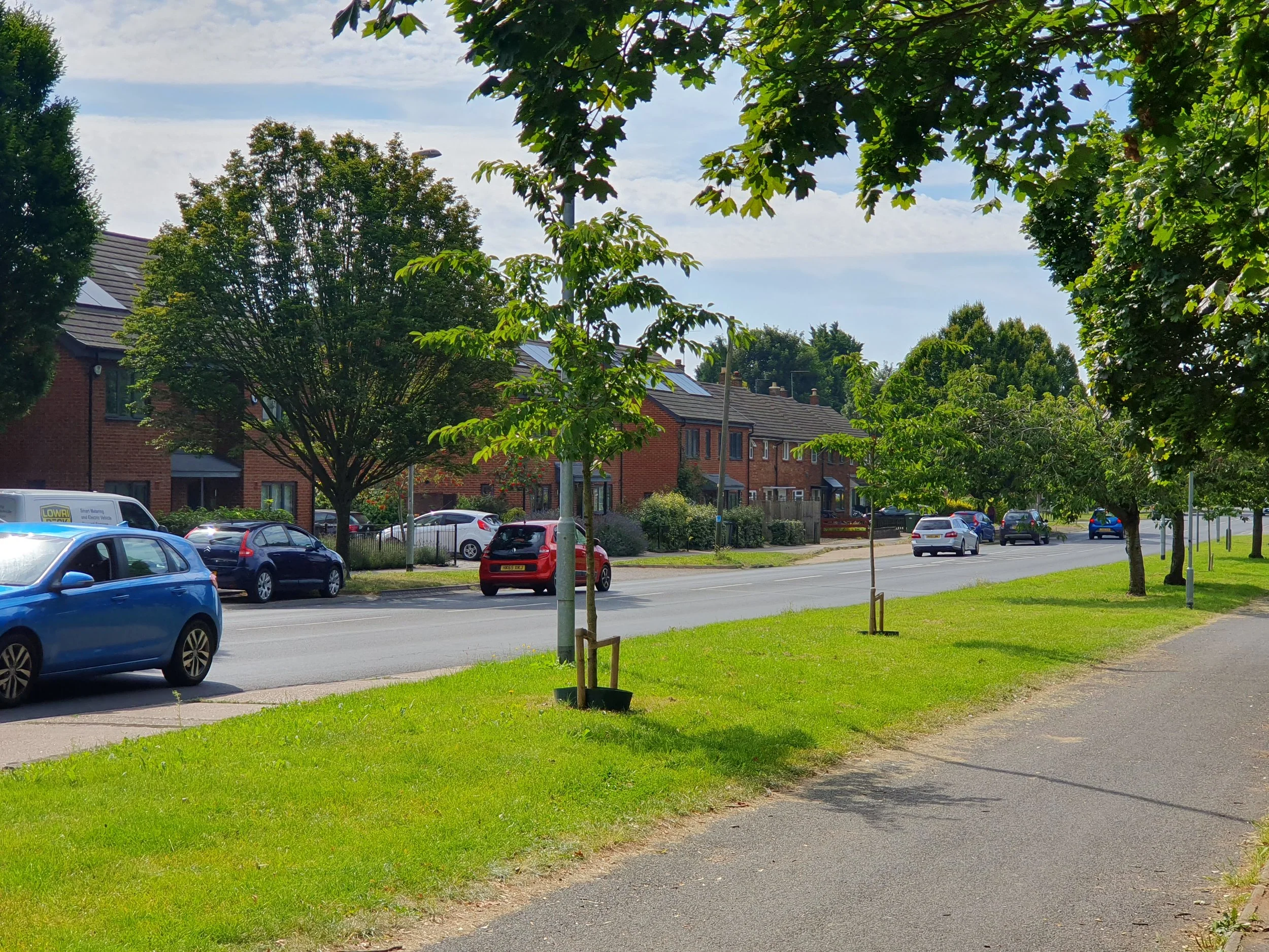 Residential street with trees, parked cars, houses, and a grassy sidewalk under a partly cloudy sky.