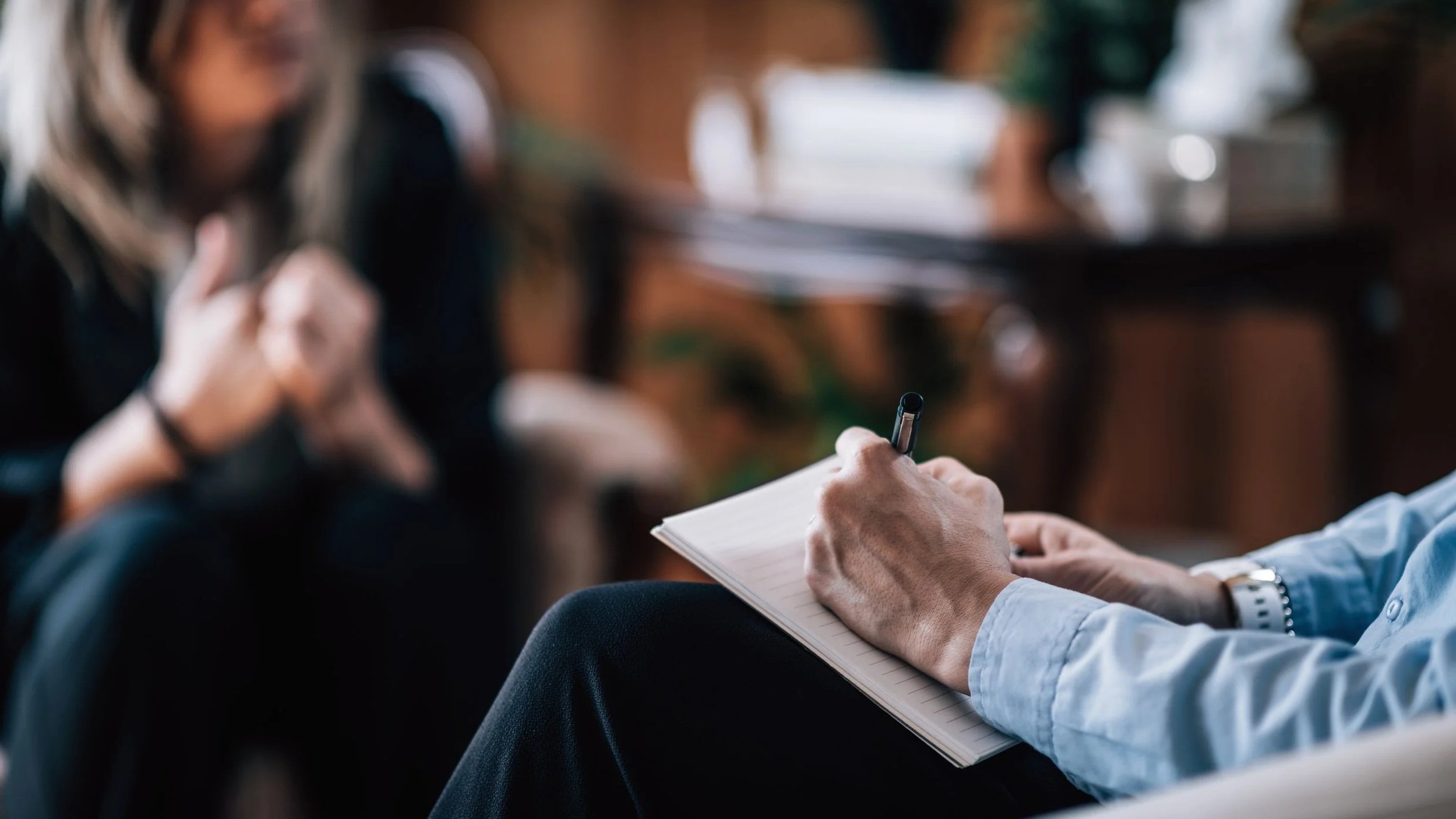 A person is sitting with a notepad and pen, likely taking notes during a therapy or counseling session, with another person in the background appearing to be a therapist.