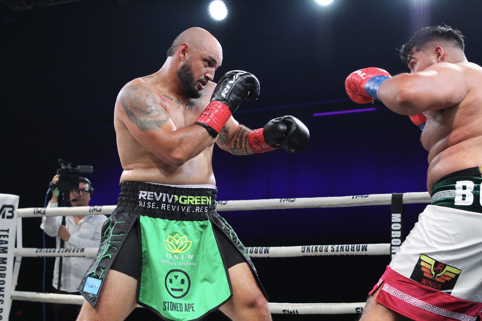 Two male boxers in a ring, one wearing green shorts with black and green design, the other wearing white shorts with red and black design, engaged in a match with the boxer on the left preparing to throw a punch.