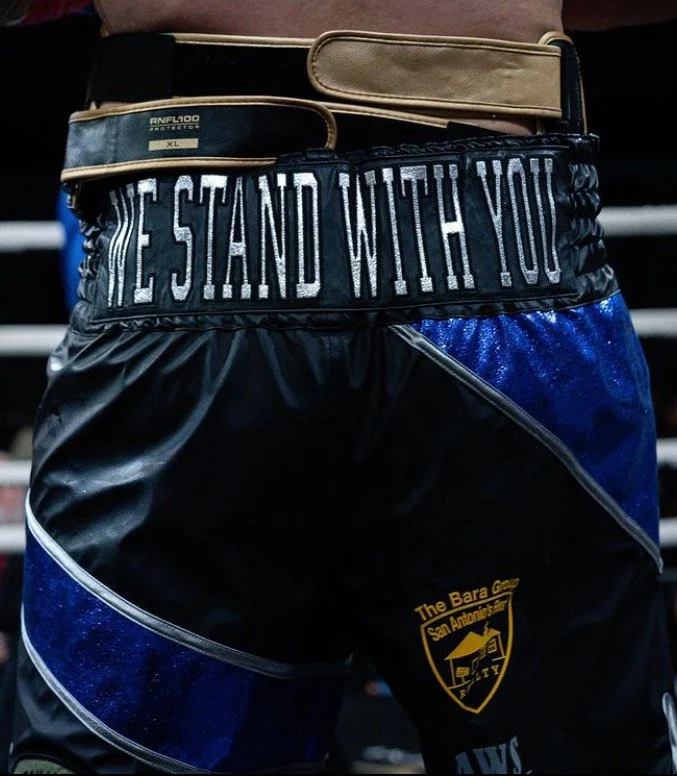 Close-up of a person's waist wearing boxing shorts with an elastic waistband that says 'WE STAND WITH YOU', and a belt with a tan pouch.