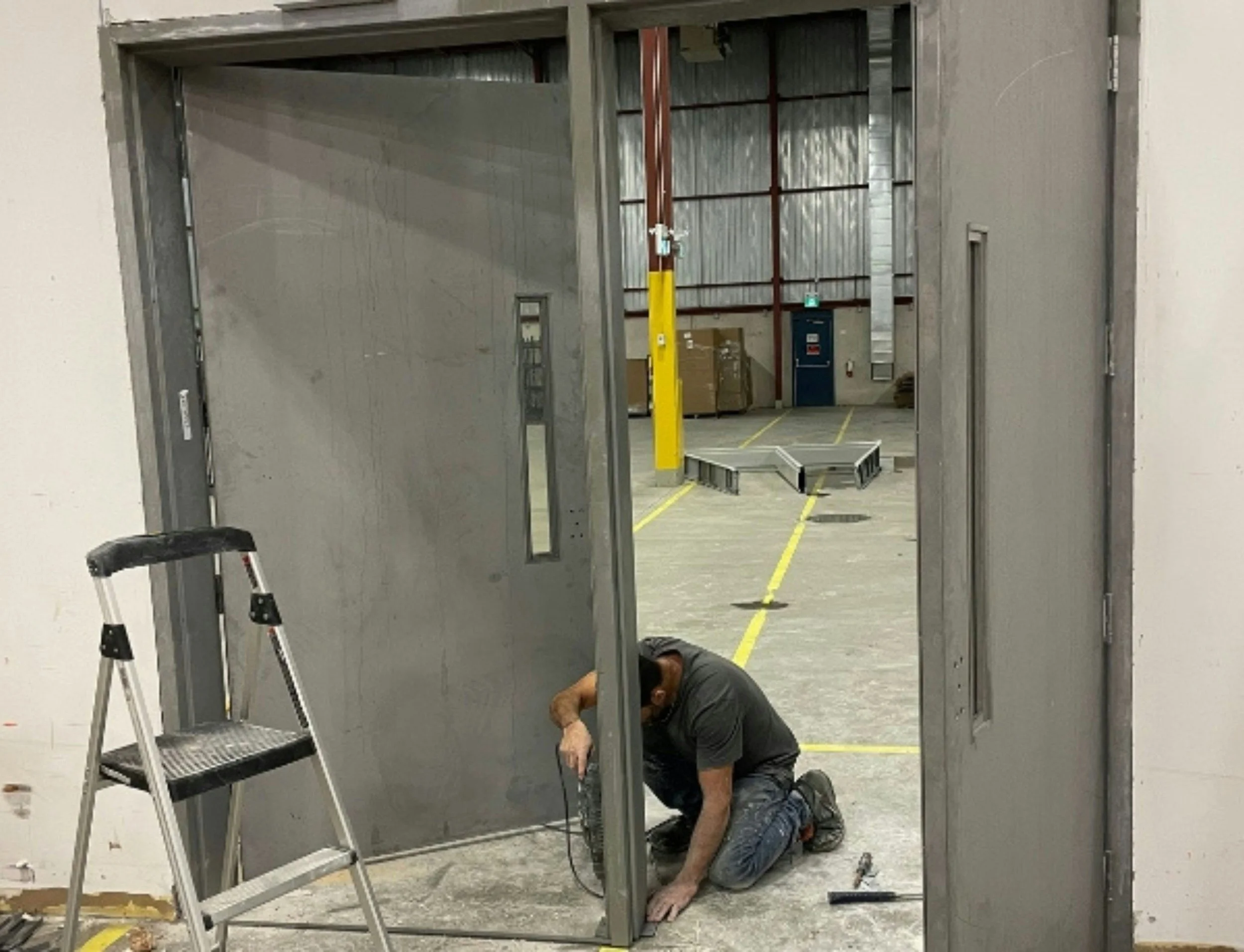 A worker kneeling on the floor installing a metal door frame inside a large industrial warehouse. A small step ladder is positioned next to the worker, and the open door frame reveals the spacious warehouse interior with yellow lines on the concrete floor and industrial equipment in the background.