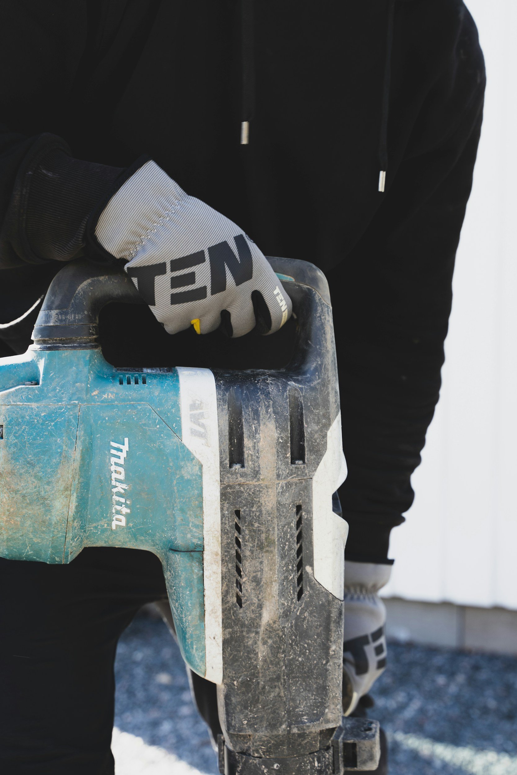 A person wearing gray work gloves holding a blue cordless power saw, with a black hoodie and black pants, standing outside on gravel.