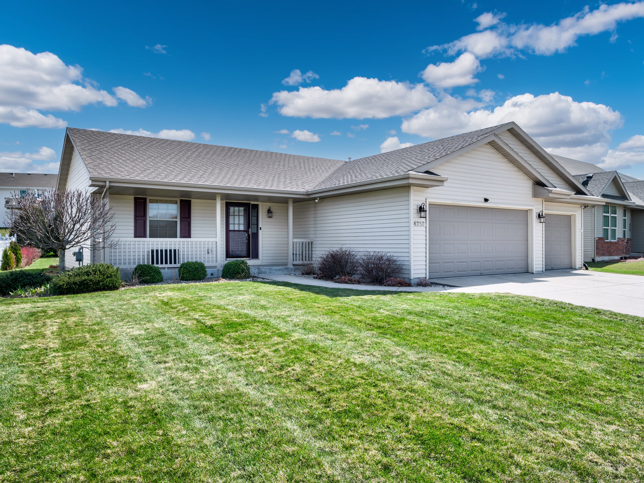 Single-story white house with a three-car garage, front porch, and manicured front yard on a sunny day with blue sky and fluffy clouds.