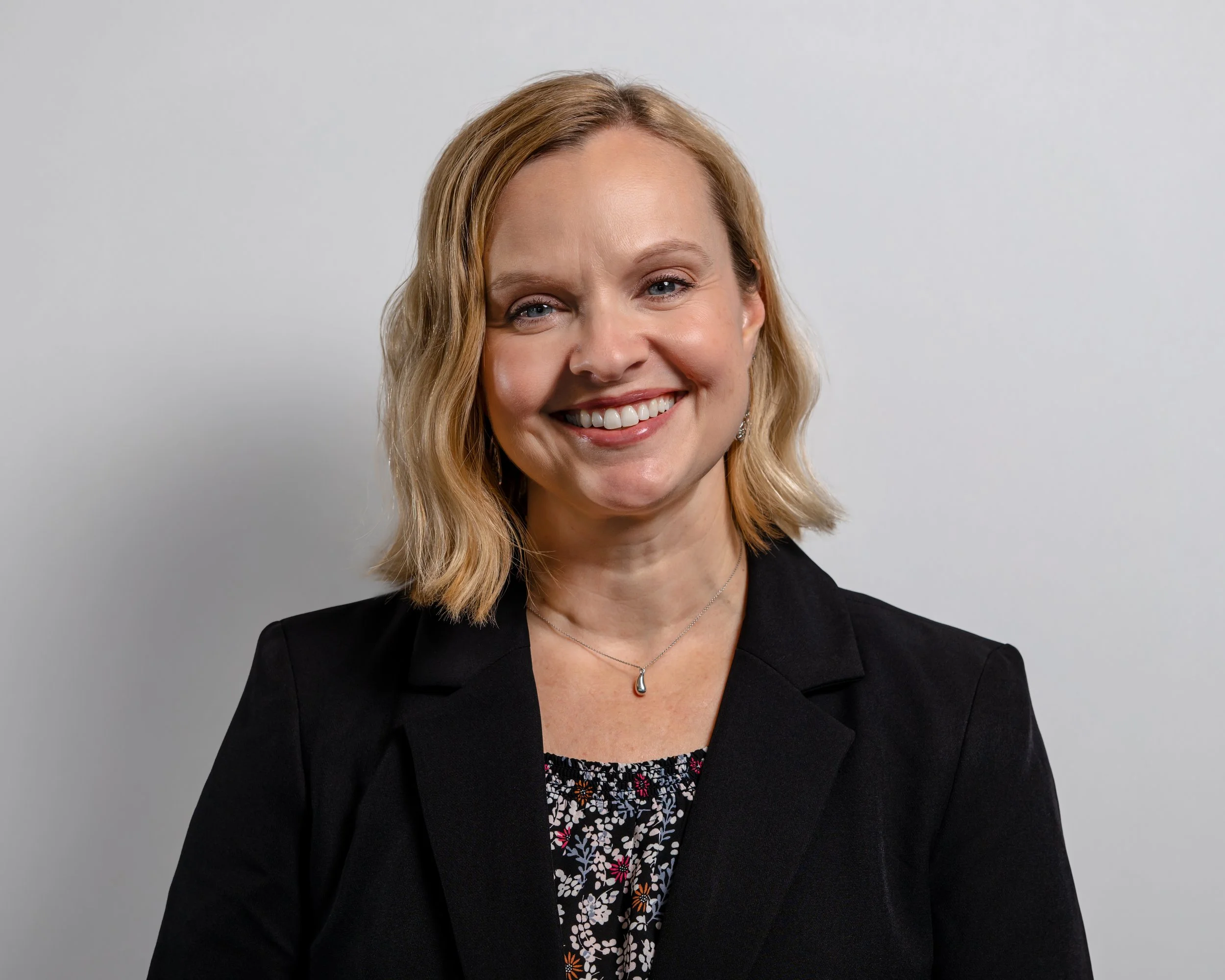 A woman with shoulder-length blonde hair, smiling, wearing a black blazer and a floral top, standing against a plain light gray background.