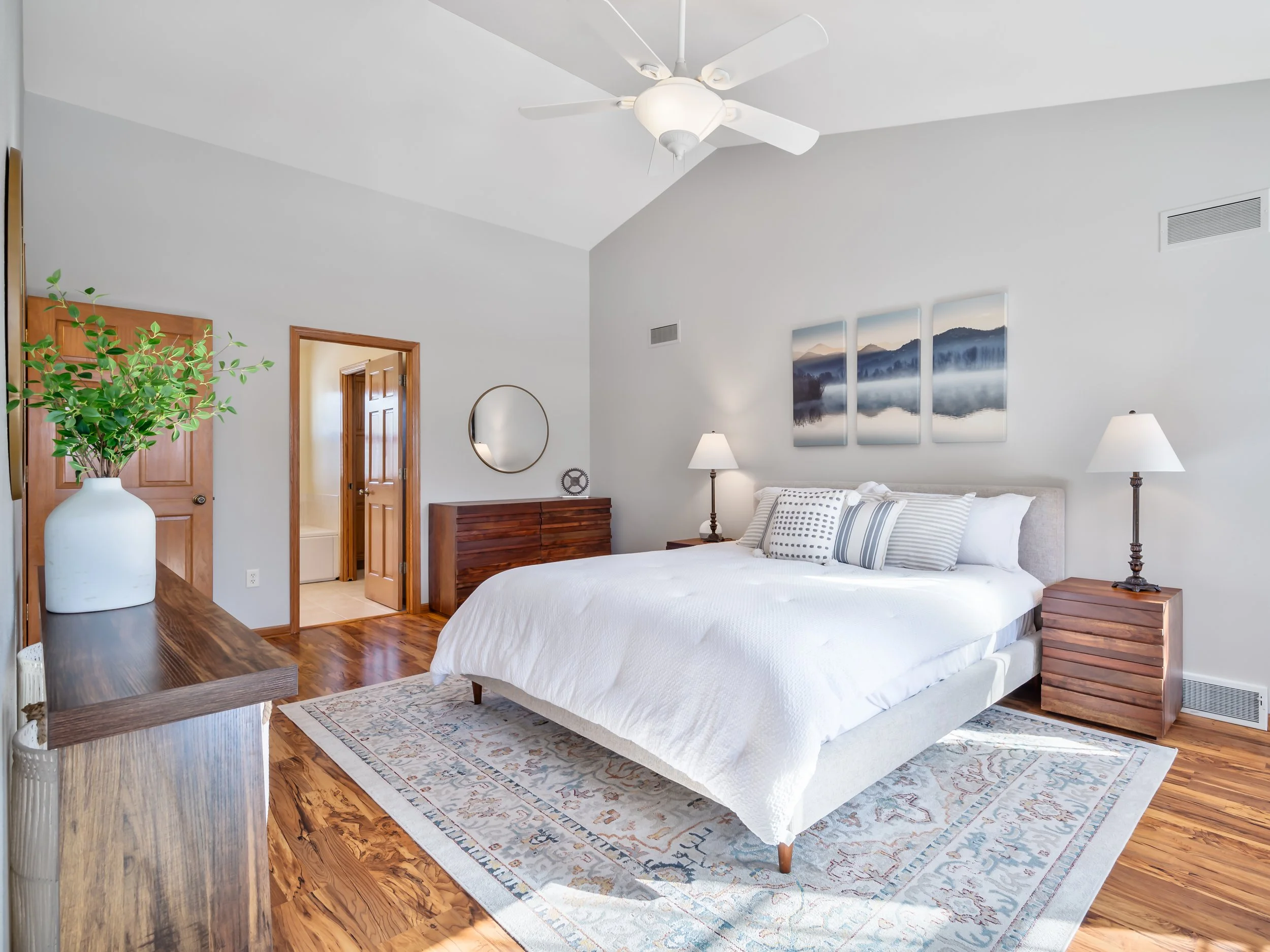 A bedroom with white walls, a bed with white bedding and pillows, wooden bedside tables and lamps, a wooden dresser, a decorative mirror, a large potted plant, and landscape artwork above the bed.