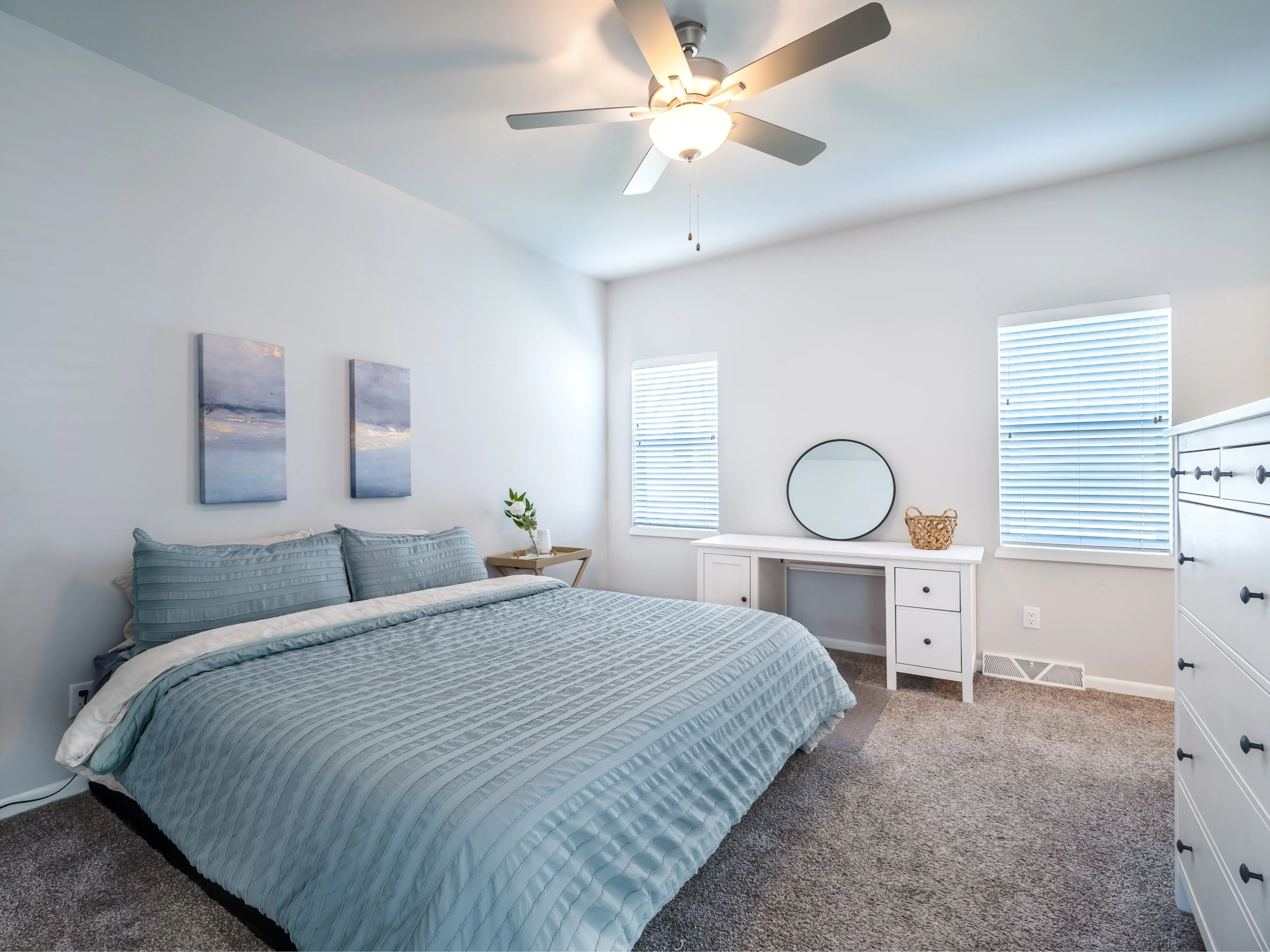 A bedroom with white walls and ceiling, a ceiling fan with lights, a bed with blue striped bedding, two framed pictures of a sky over water above the bed, a small wooden side table with a plant, a white desk with a round mirror, a wicker basket, and 