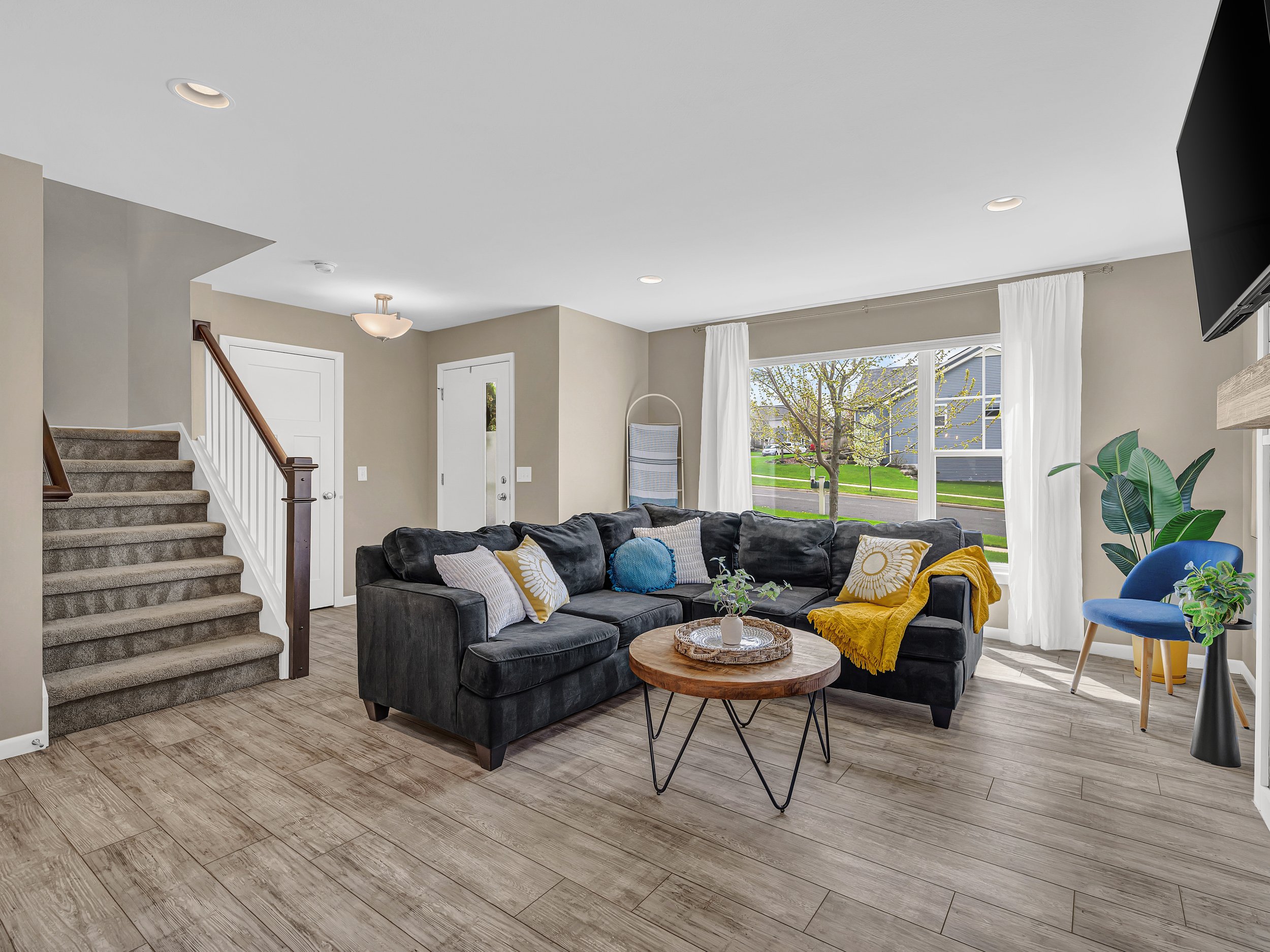 Living room with beige walls, large window with white curtains, black sectional sofa with decorative pillows, round wooden coffee table with potted plant, blue accent chair, staircase with carpeted steps, hardwood flooring.