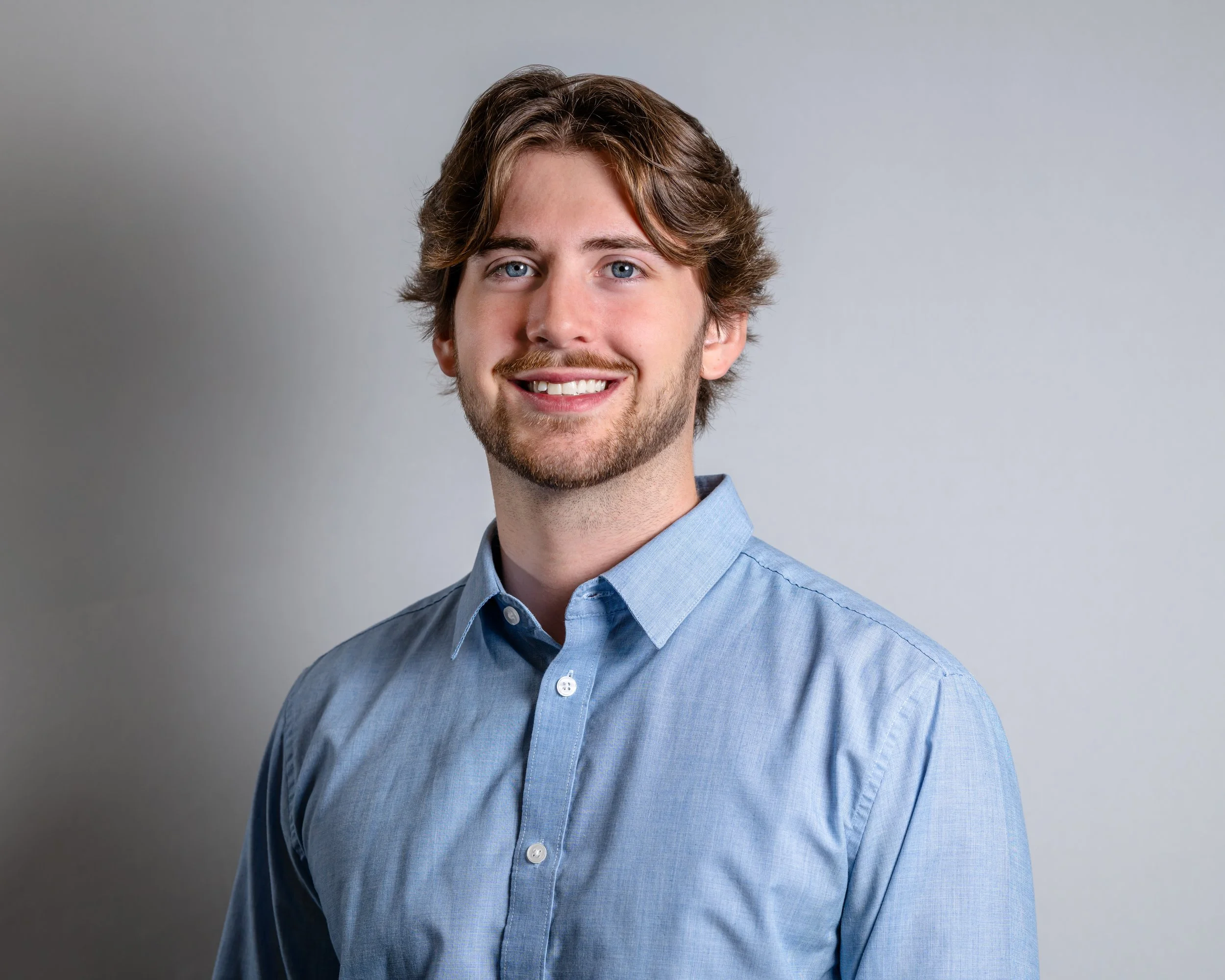A young man with brown hair, blue eyes, and a beard, wearing a light blue button-up shirt, smiling against a plain light gray background.