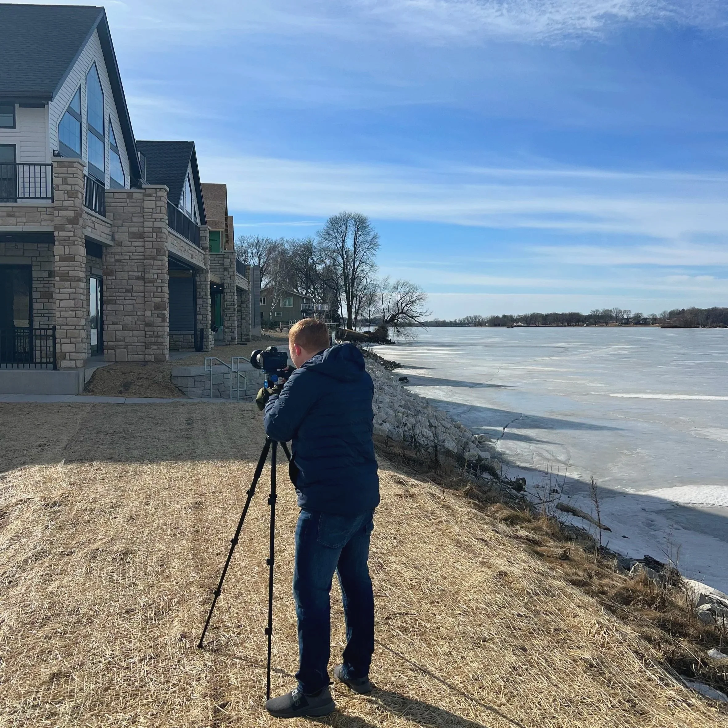 A person operating a camera on a tripod by a frozen lake with houses and leafless trees in the background on a clear, sunny day.