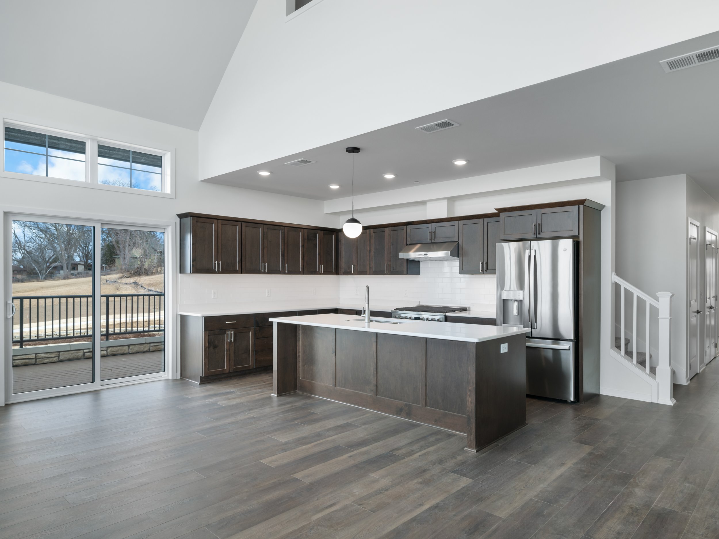 Modern kitchen with dark wood cabinets, stainless steel refrigerator, island with white countertop, and large sliding glass door leading to outdoor patio.