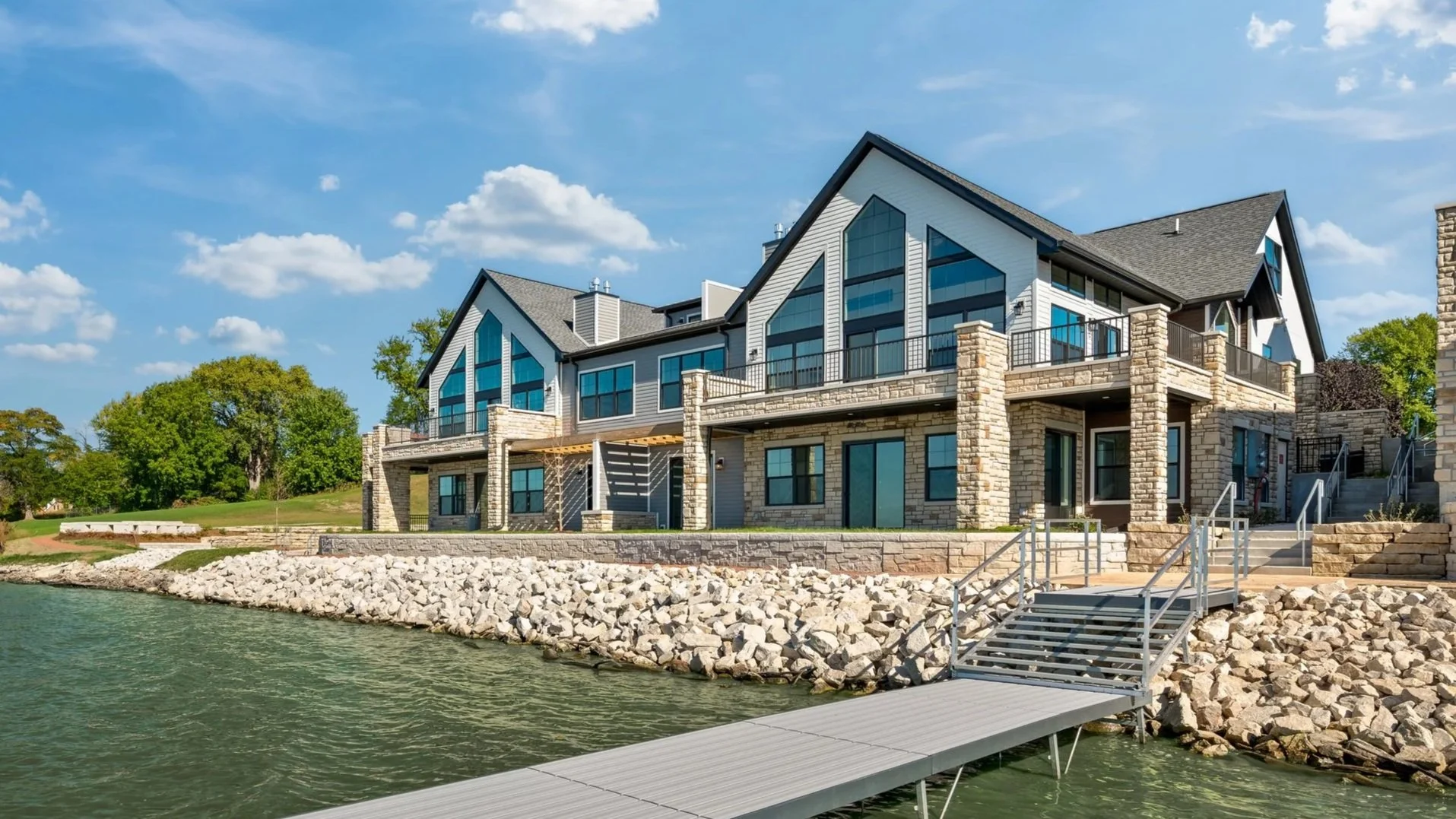 Large modern house with multiple levels and large triangular windows by the water, with a dock leading to the water and surrounded by trees and greenery.