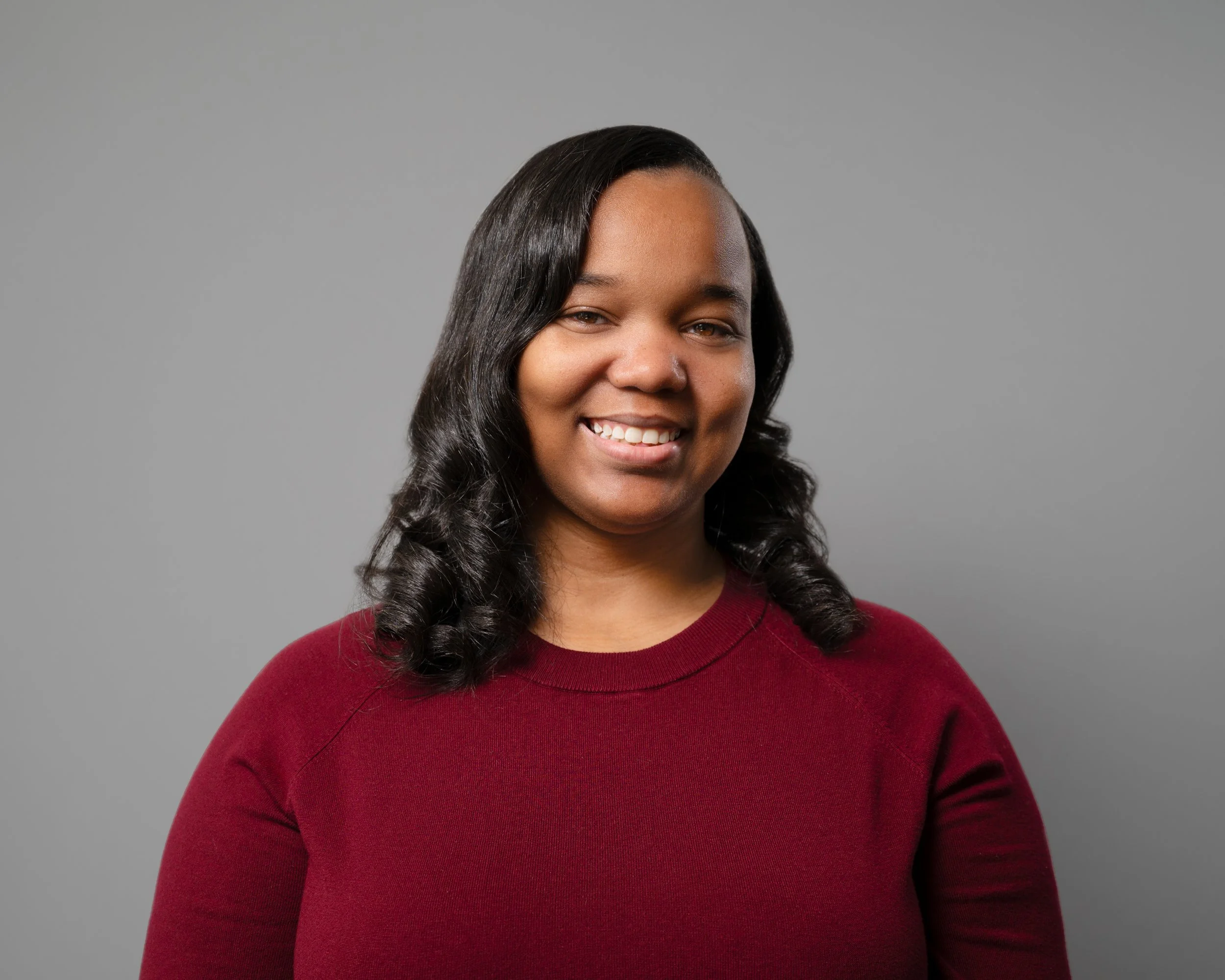 A woman with dark, curly hair wearing a red sweater smiles in front of a plain gray background.
