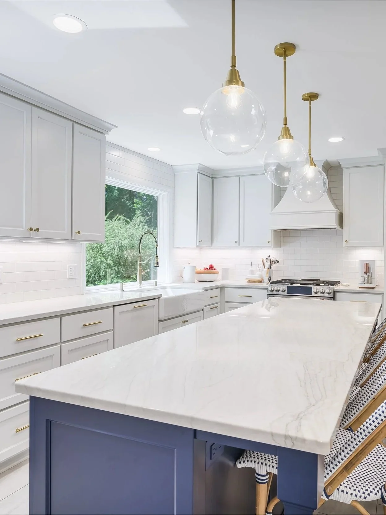 Bright white kitchen with gold and glass pendant lights, white cabinets, and a large marble island with chairs. A window overlooks greenery.