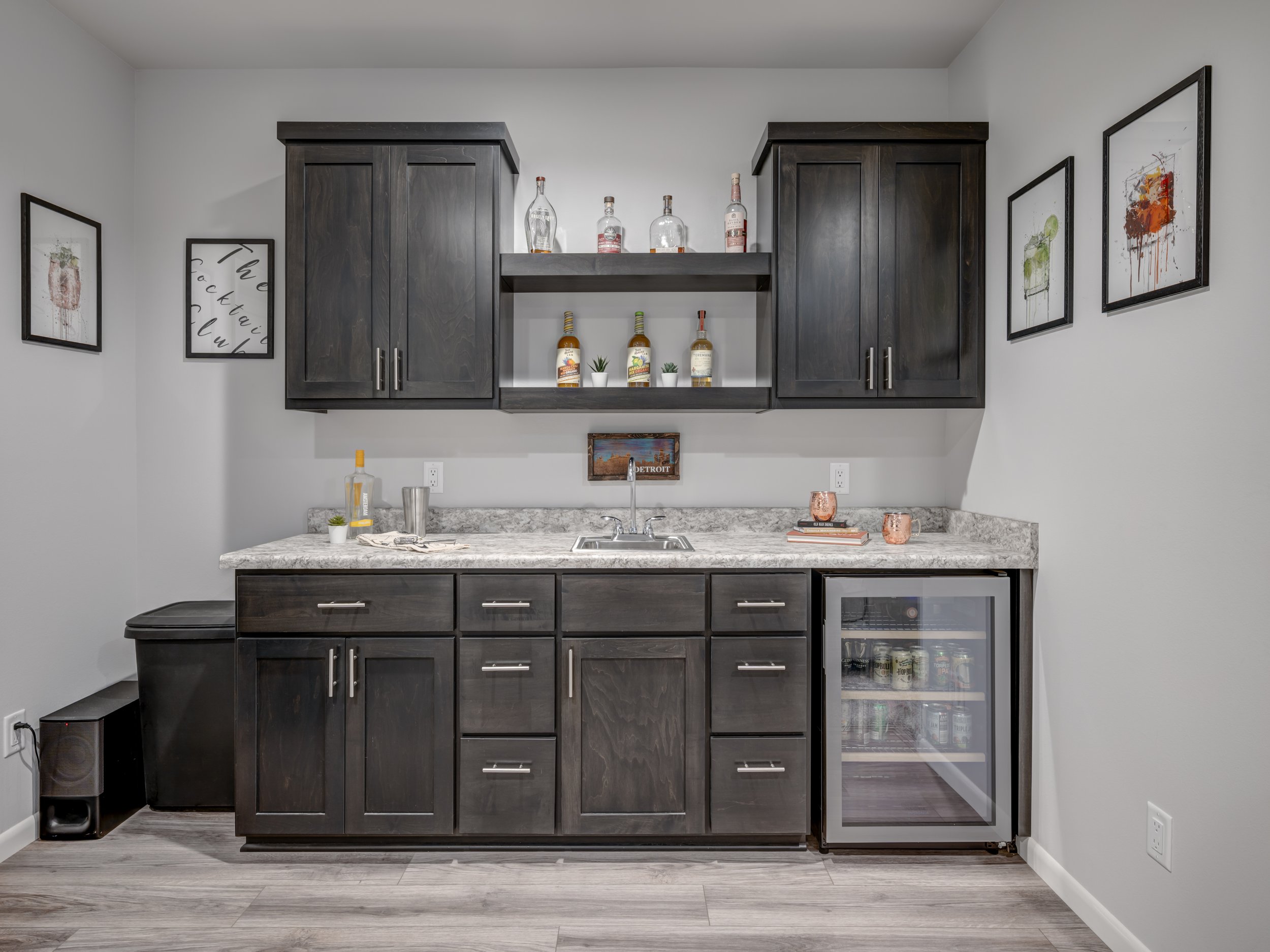 A small kitchen wet bar with dark wood cabinets, a granite countertop, and a beverage fridge with drinks inside. Decor includes framed art on the walls and bottles of alcohol on shelves.
