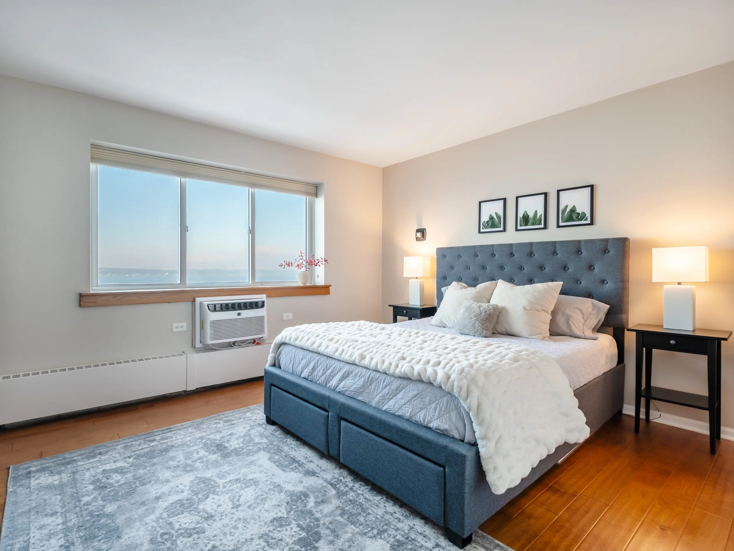 A bedroom with a large window, gray bed with pillows, nightstands with lamps, and framed artwork on the wall