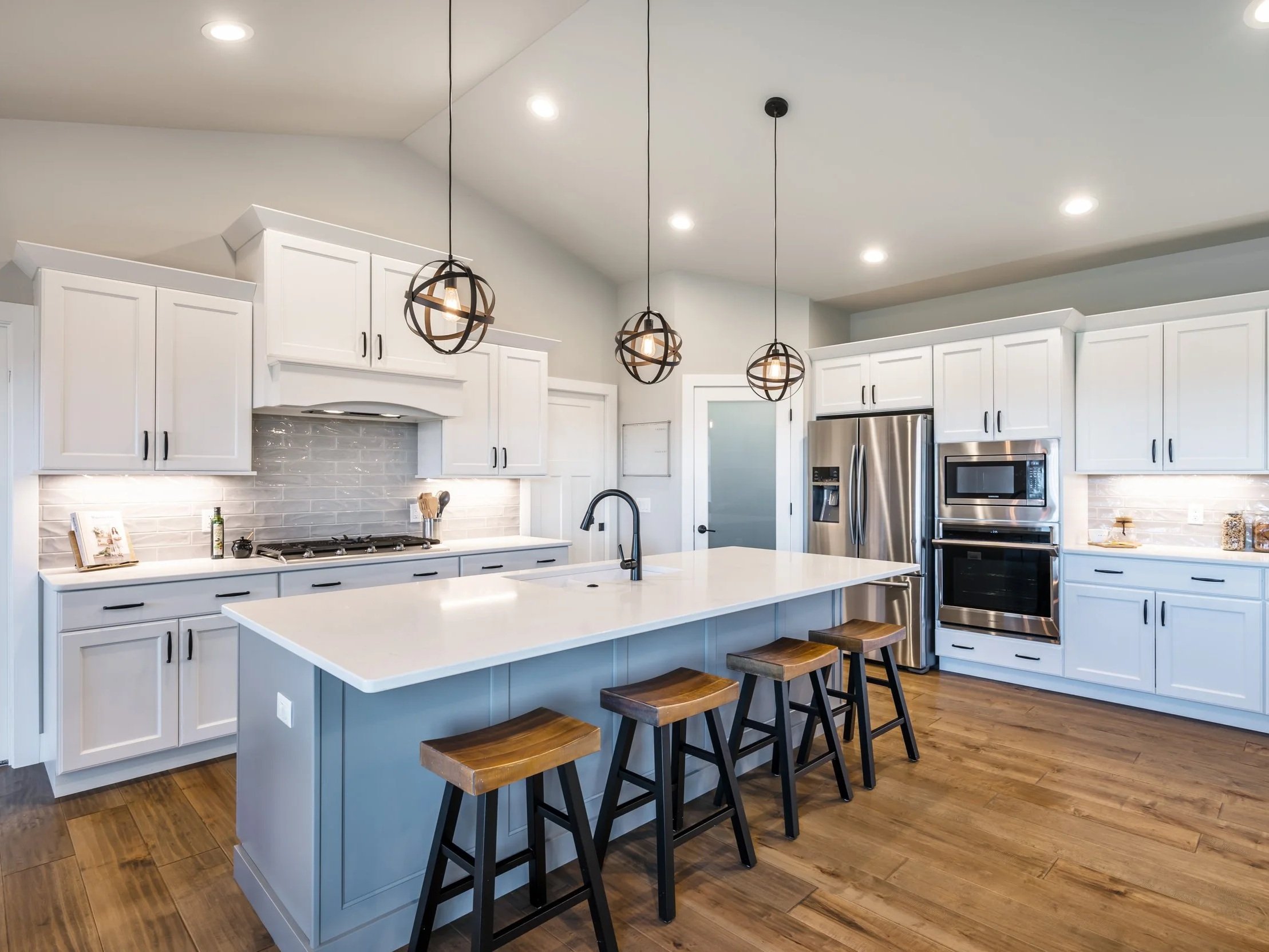Modern kitchen with white cabinets, stainless steel refrigerator, oven, microwave, and a large island with bar stools, pendant lighting, hardwood floors.