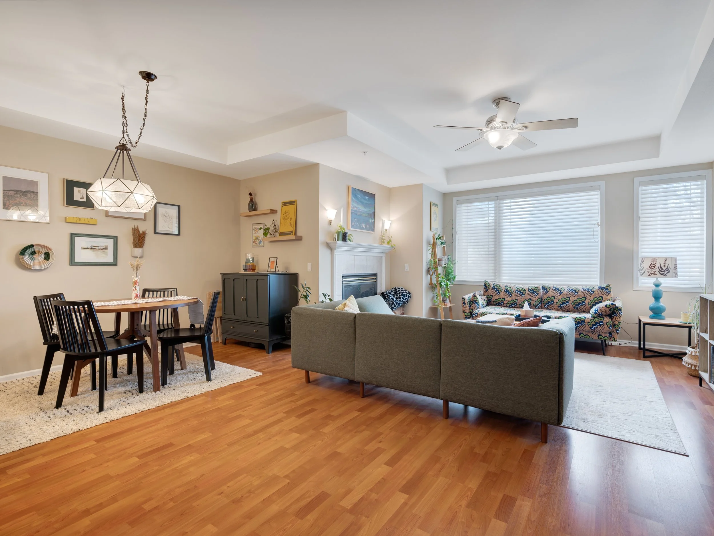 Living room with wooden floor, beige walls, a gray sofa, a patterned armchair, a white rug, a black entertainment cabinet, a fireplace with artwork above, a window with blinds, and various decorative items including a blue lamp and plants.