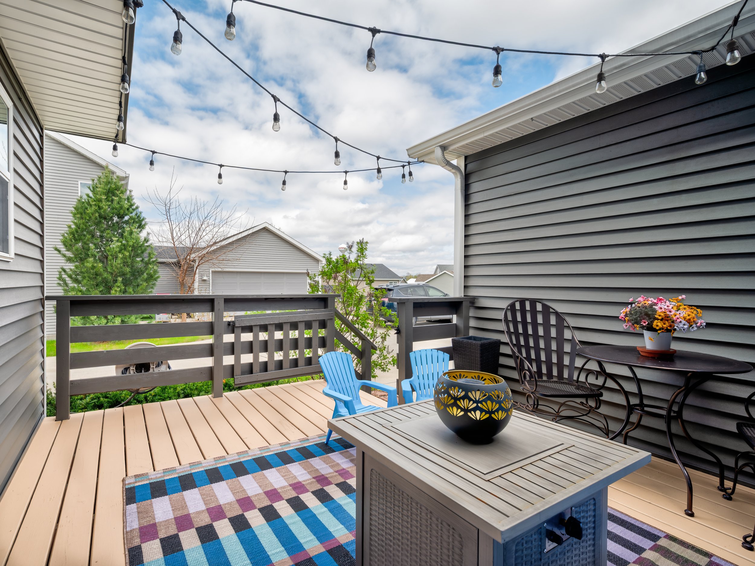 Outdoor deck area with string lights, a small table with a black bowl, a flowerpot with colorful flowers, a black metal chair, a small blue plastic chair, and a checkered rug, surrounded by houses with gray siding and a partly cloudy sky.