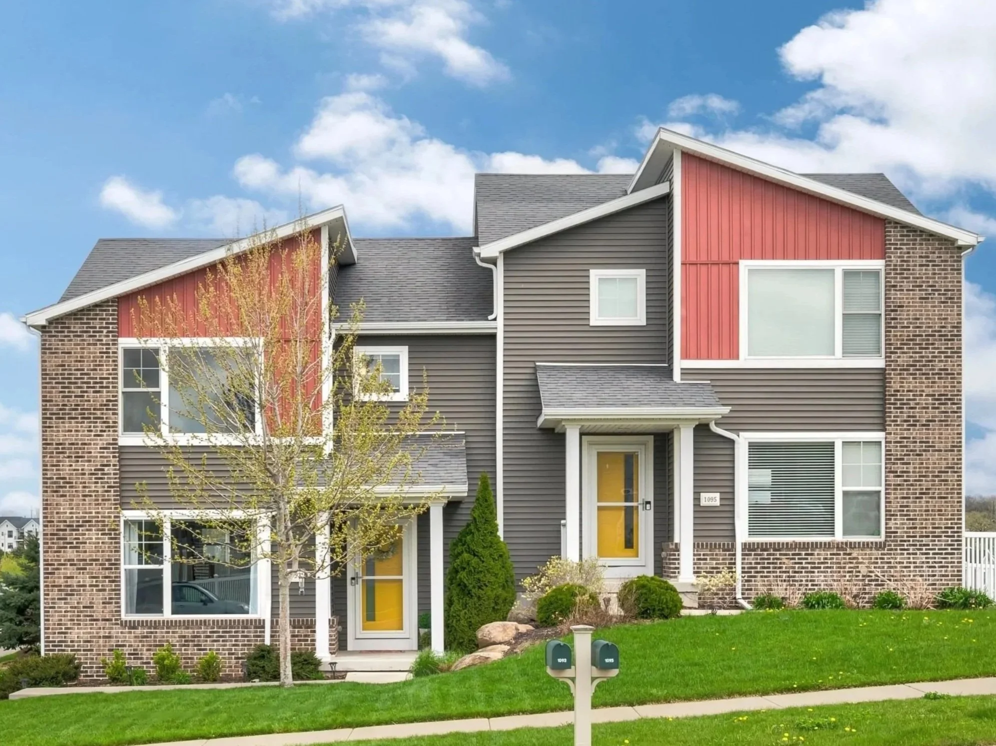 A modern two-story house with a mix of brick and gray siding, decorative red panels, and yellow doors, surrounded by a well-kept lawn and a young tree.