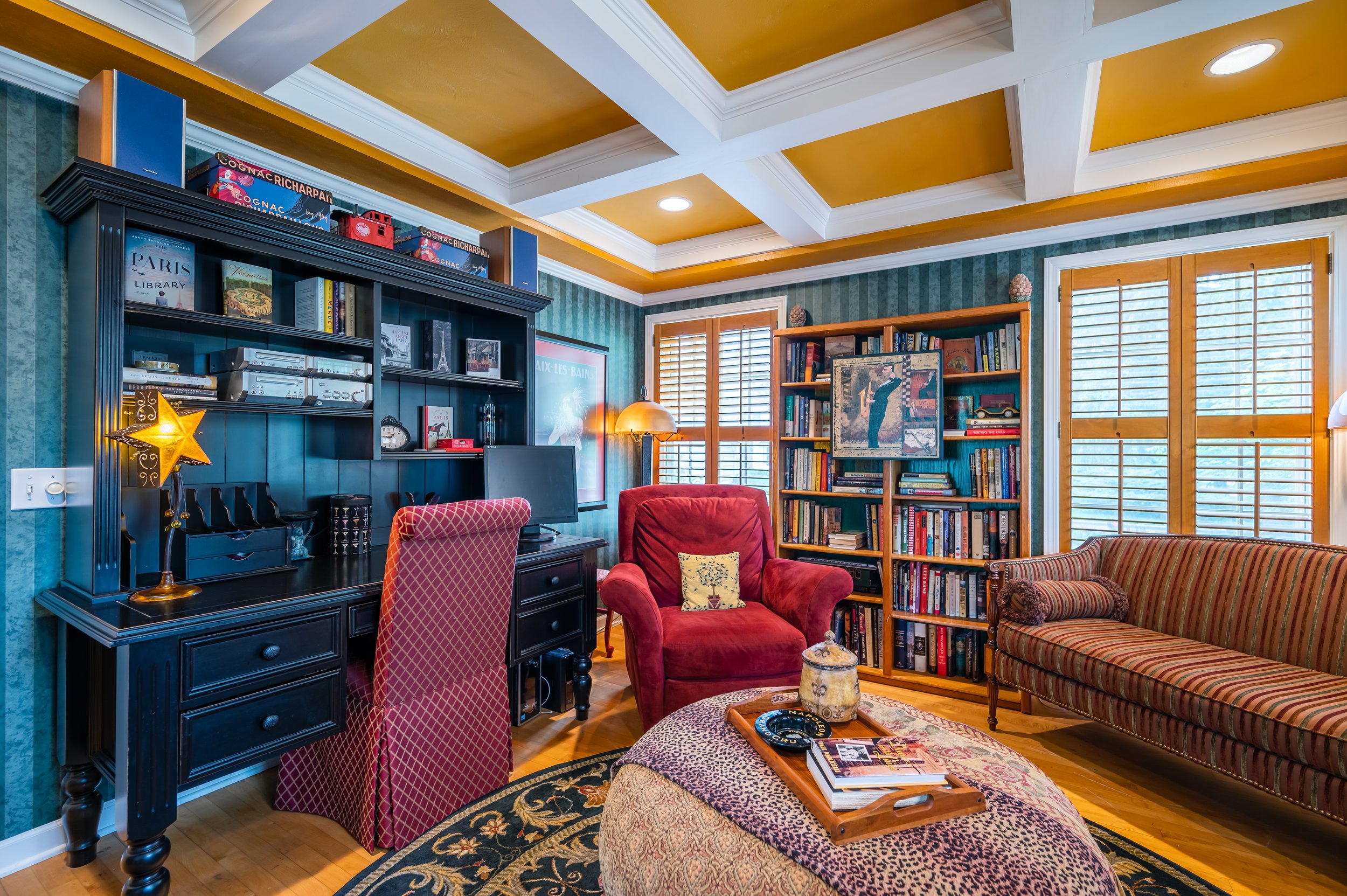 A cozy living room with green striped wallpaper, wooden trim, and a coffered ceiling painted yellow and white. The room contains a black desk with a computer, a red armchair with a yellow pillow, a striped sofa, a wooden bookshelf filled with books, 