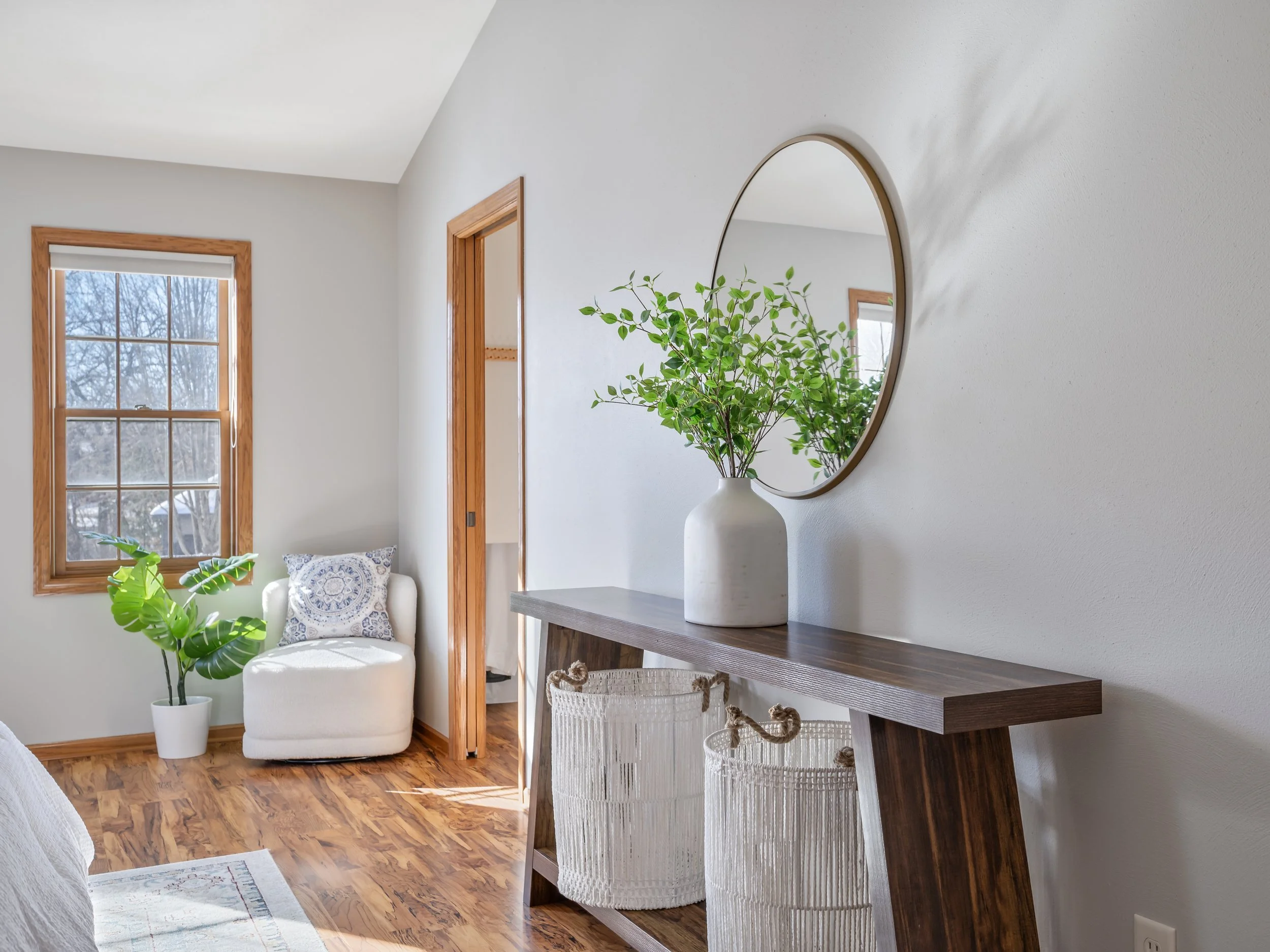 Interior of a bright, minimalist living space with a wooden floor, a white armchair with decorative pillow, a potted plant, a wall mirror, and a wooden console table with a potted green plant.