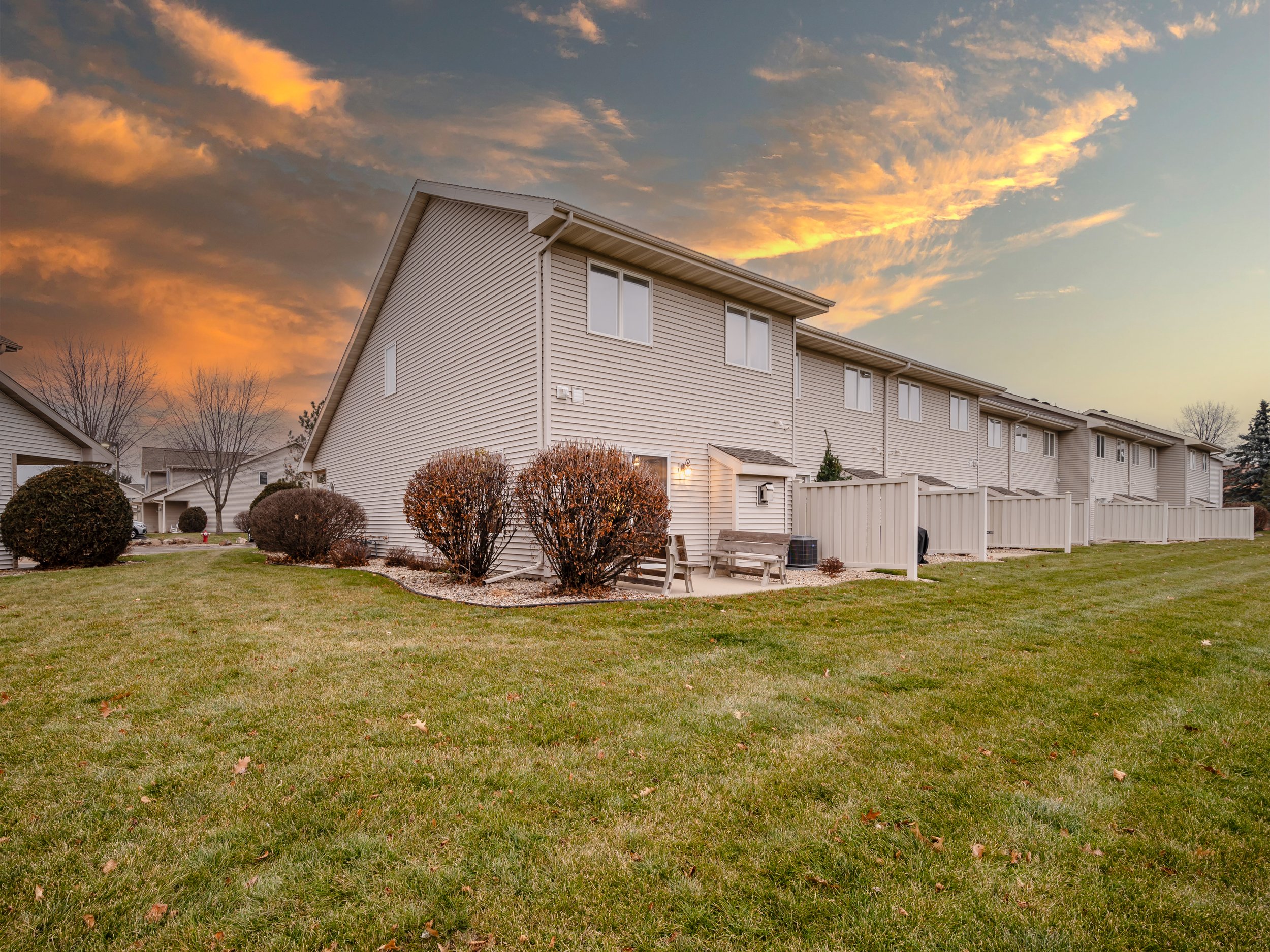 A row of beige townhouses with vinyl siding. The backyards feature well-maintained grass, bushes, a bench, and a small patio area. The sky is partly cloudy at sunset, with orange and yellow hues.