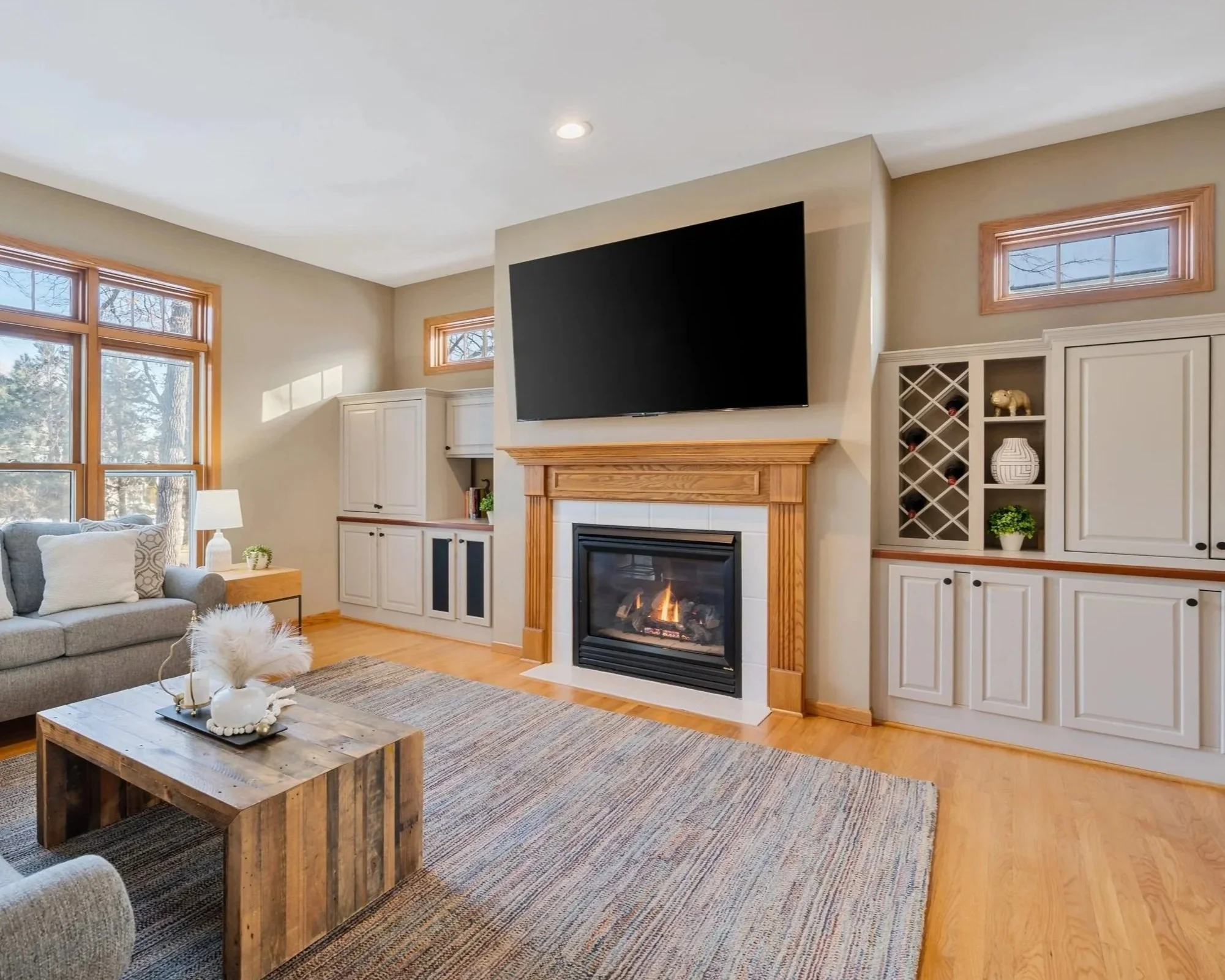 Living room with large windows, a gray sofa with pillows, a wooden coffee table, built-in white cabinets, a fireplace with a wooden mantel, and a mounted flat-screen TV above the fireplace.