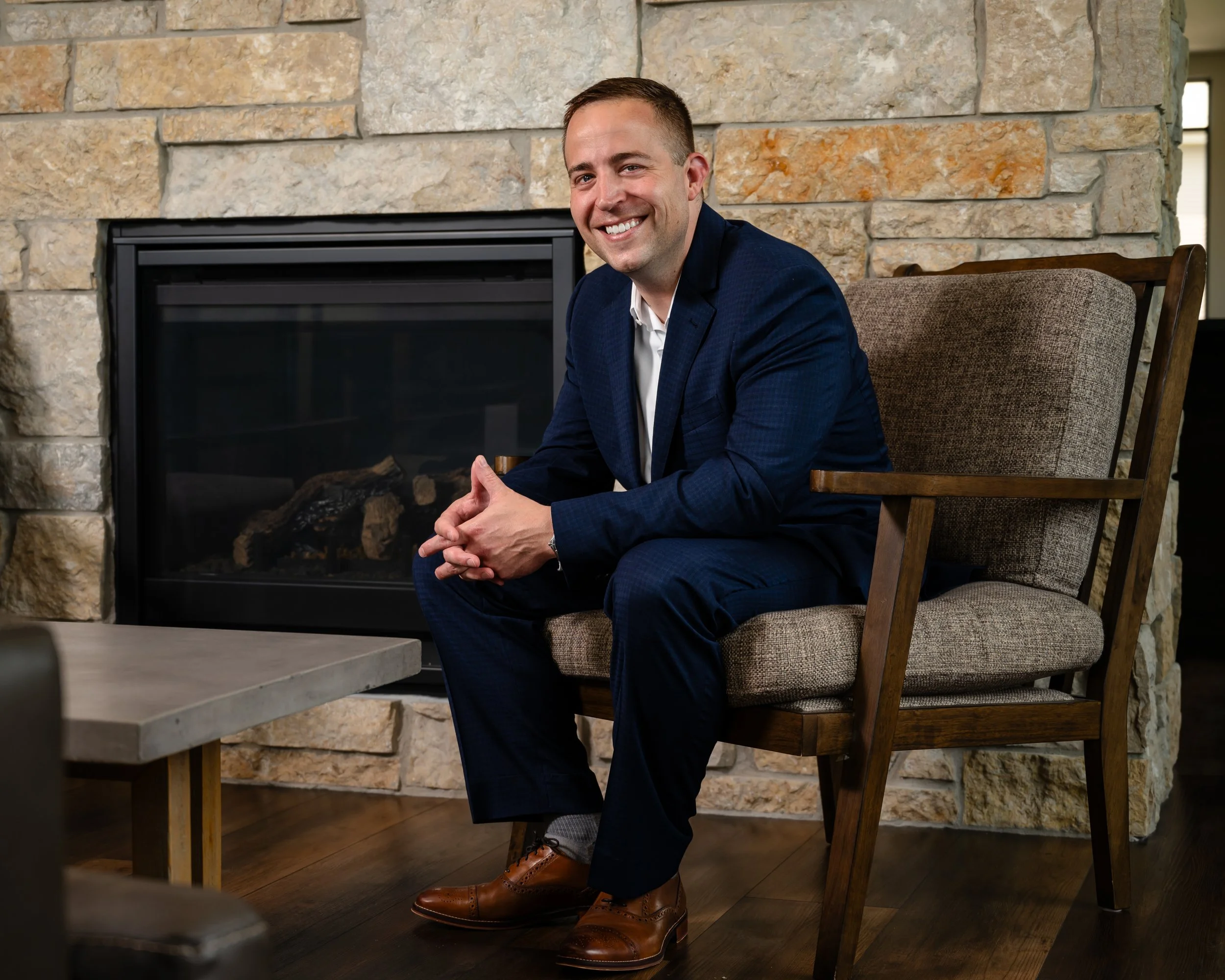 A man in a blue suit sitting on a beige chair by a fireplace, smiling and looking at the camera.
