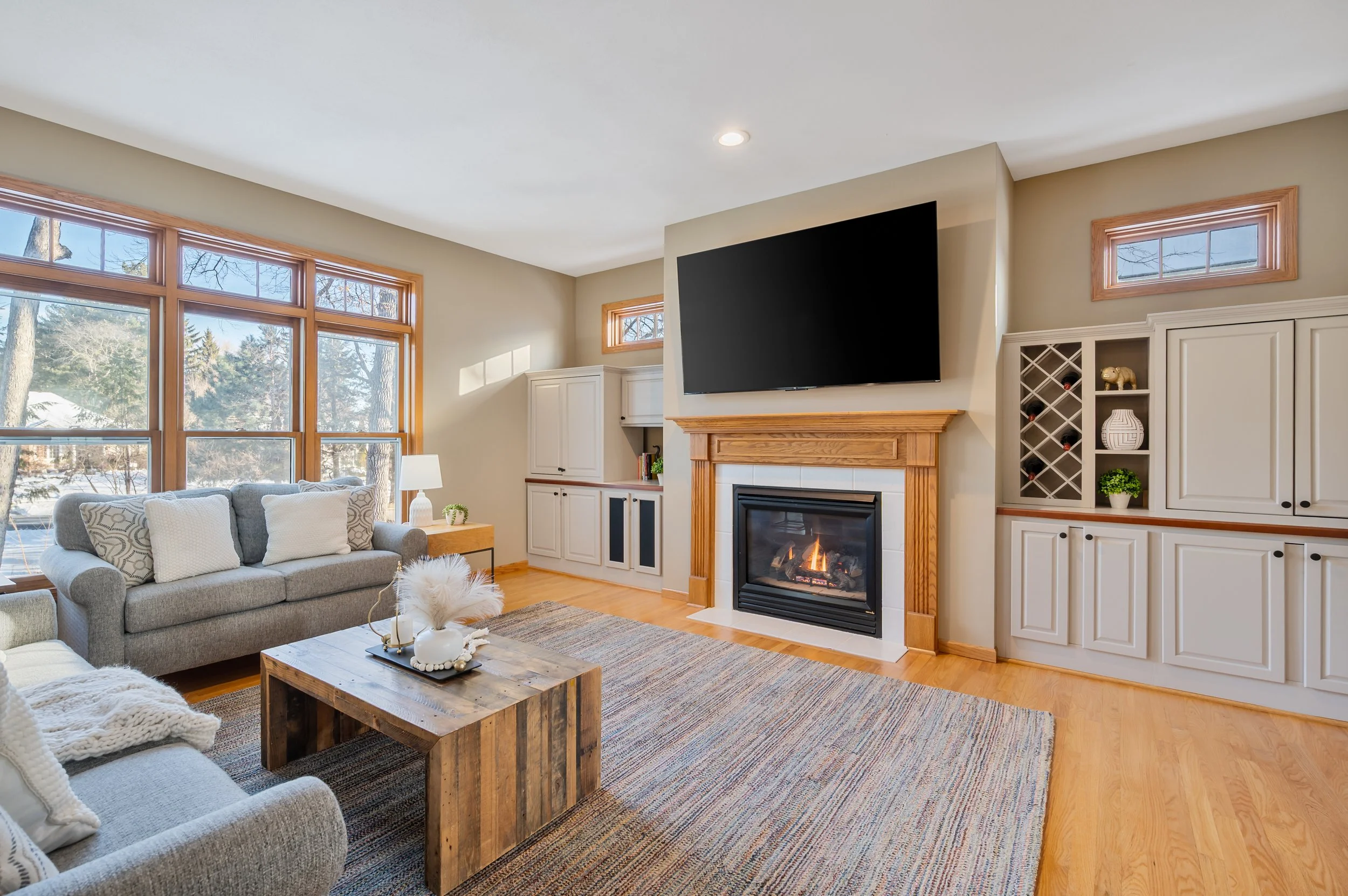Living room with large window, gray sofa, wooden coffee table, fireplace with a mounted TV, built-in cabinets, and hardwood flooring.