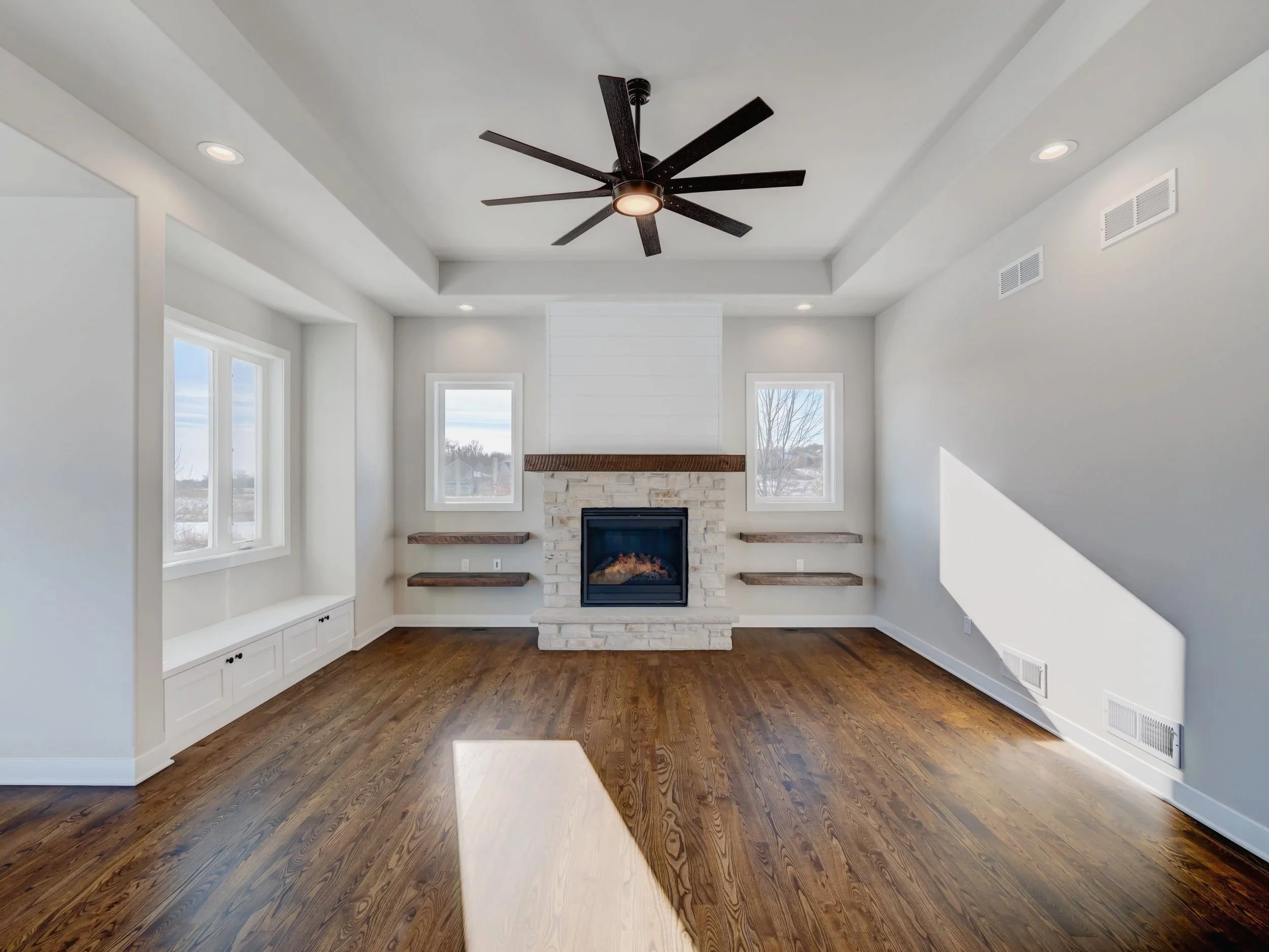 Empty living room with hardwood floors, white walls, a fireplace with stone surround, built-in shelves, two windows, and a black ceiling fan.