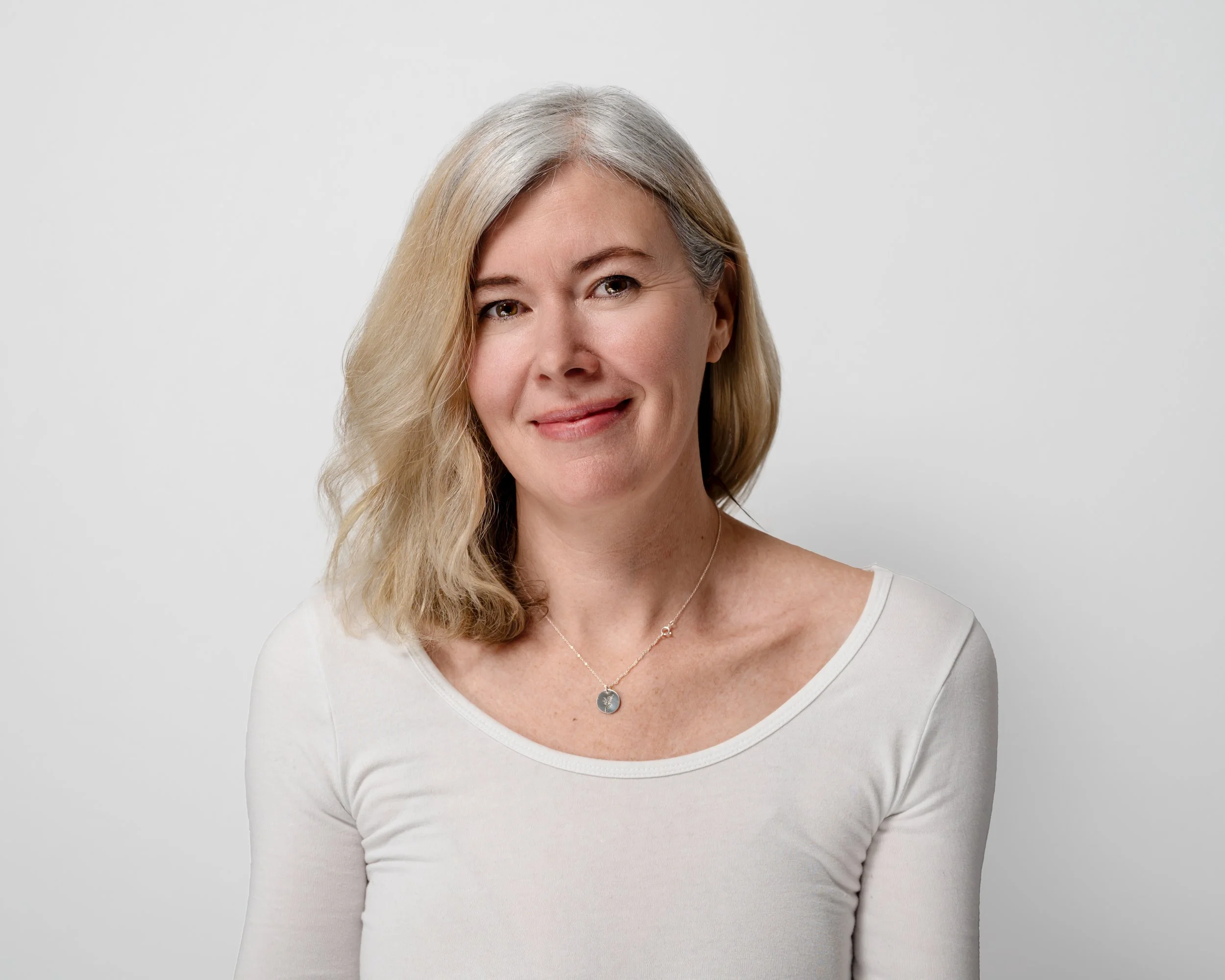A woman with shoulder-length blonde hair in a white shirt smiling against a plain white background.