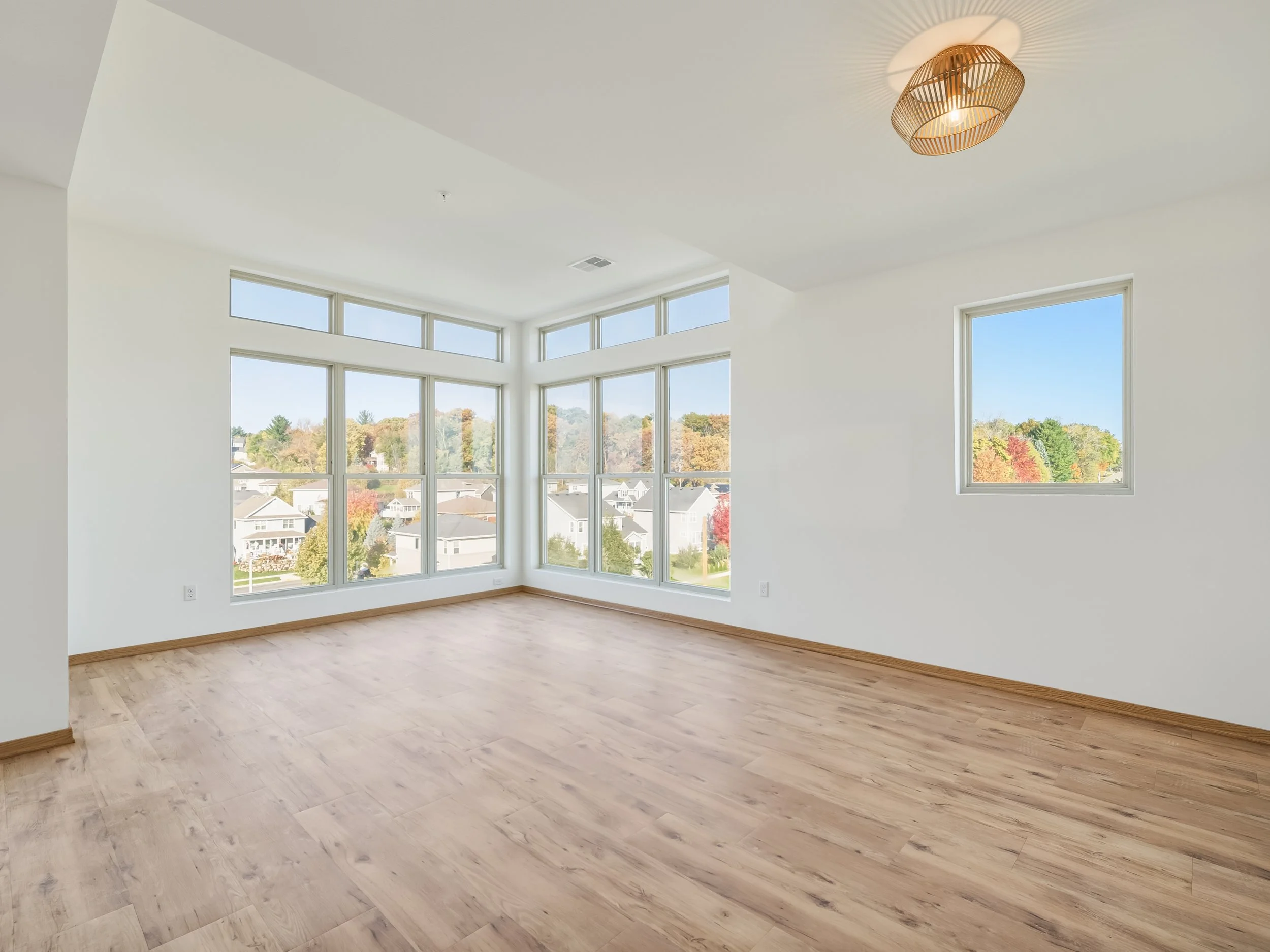 An empty room with large windows, white walls, and wooden flooring, overlooking a suburban neighborhood with trees showing fall colors.