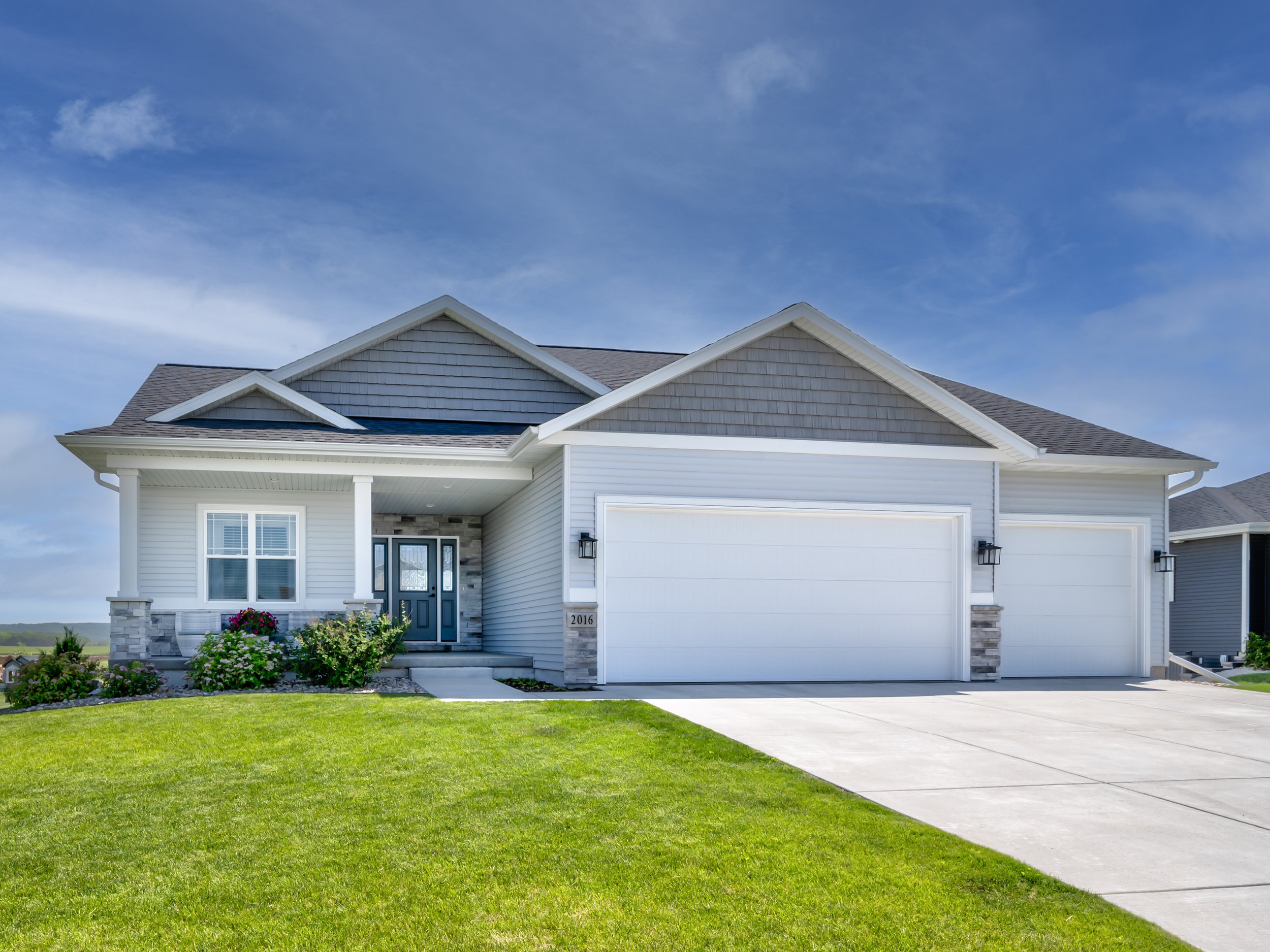 Front view of a modern, suburban house with a well-maintained lawn, white siding, gray roof, and double garage doors, under a blue sky.