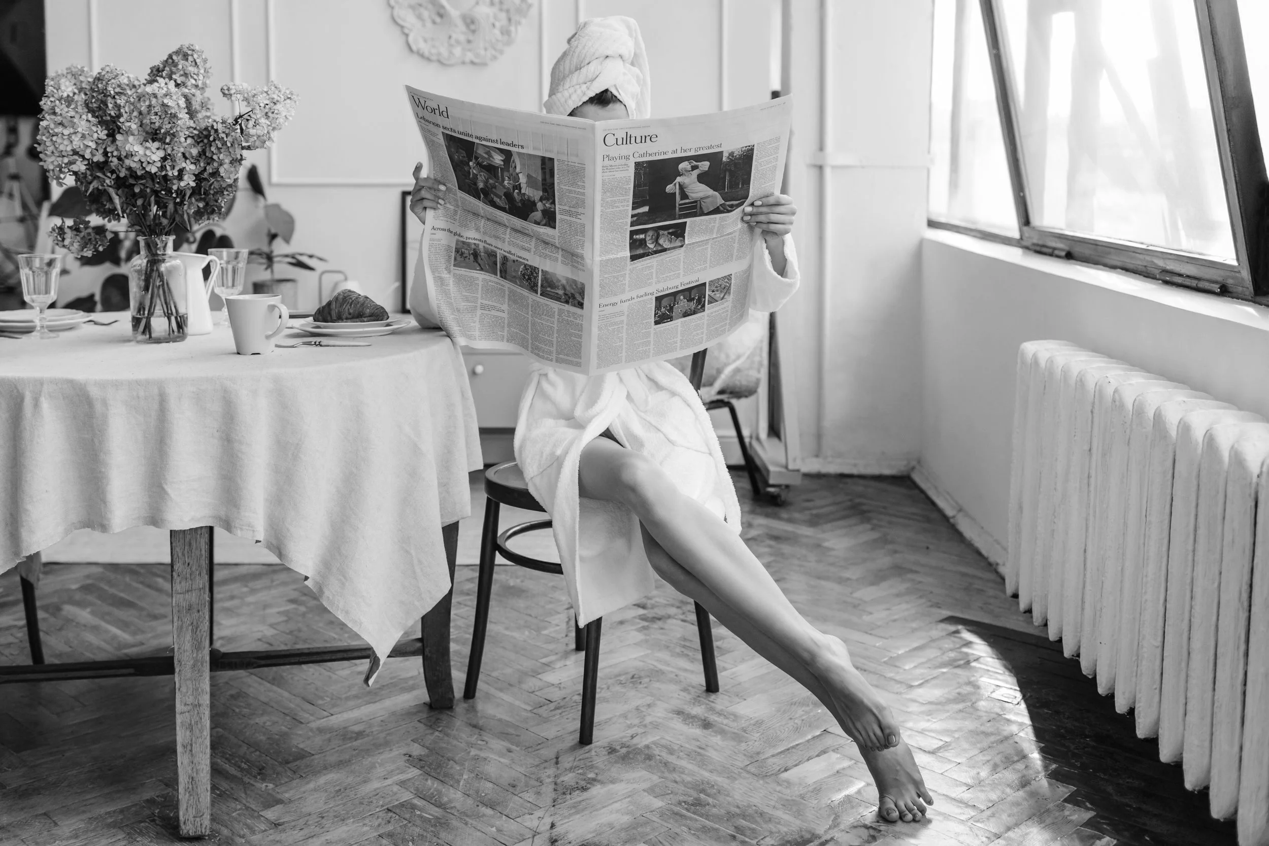 A person sitting on a stool wearing a towel on their head and a bathrobe, reading a newspaper in a bright room with a table set for breakfast and a large window.
