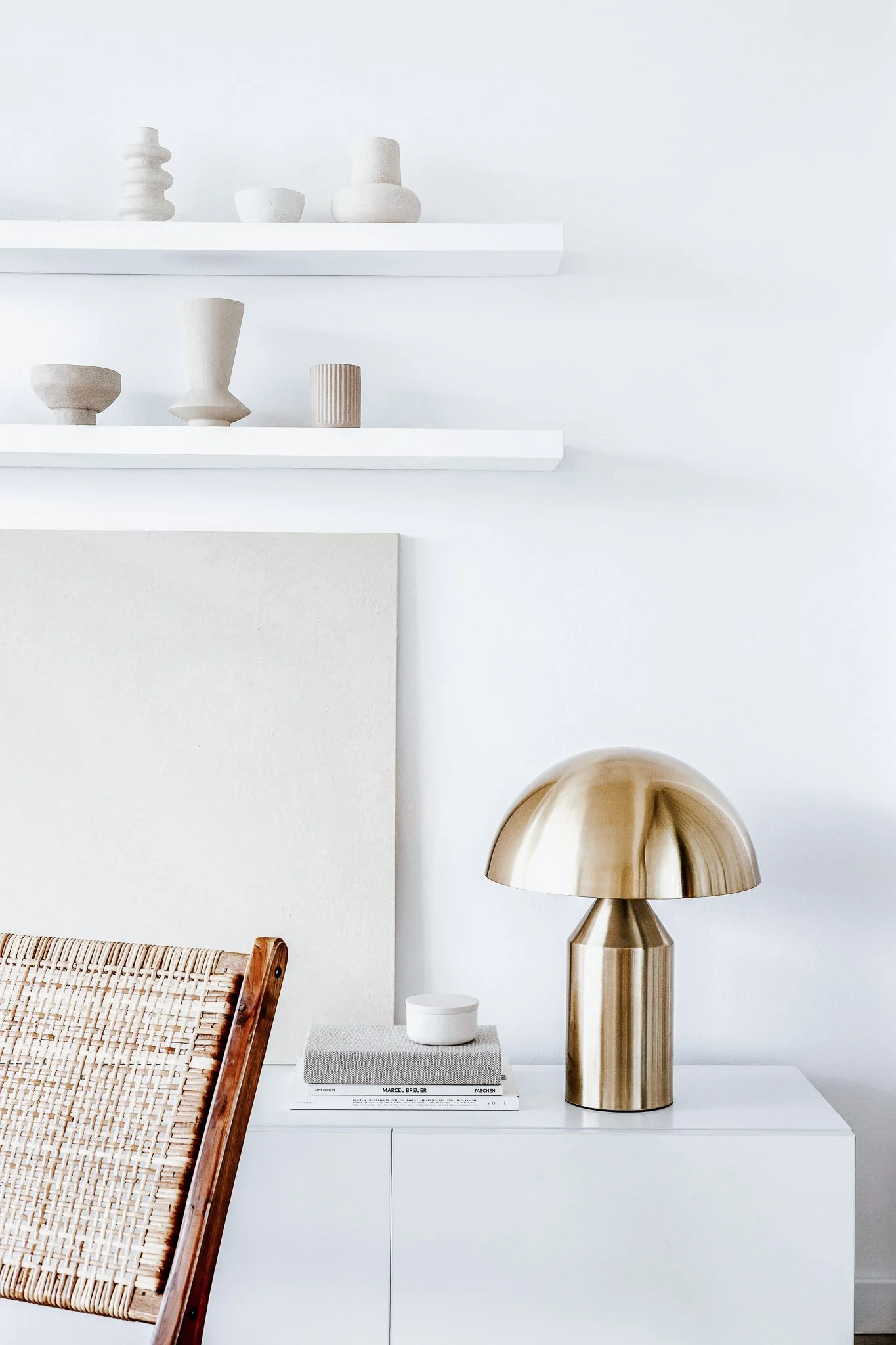 Minimalist interior scene with white shelves holding beige and white decorative vases and bowls, a white cabinet with a stack of books and a gold table lamp, and a woven chair with wooden frame.