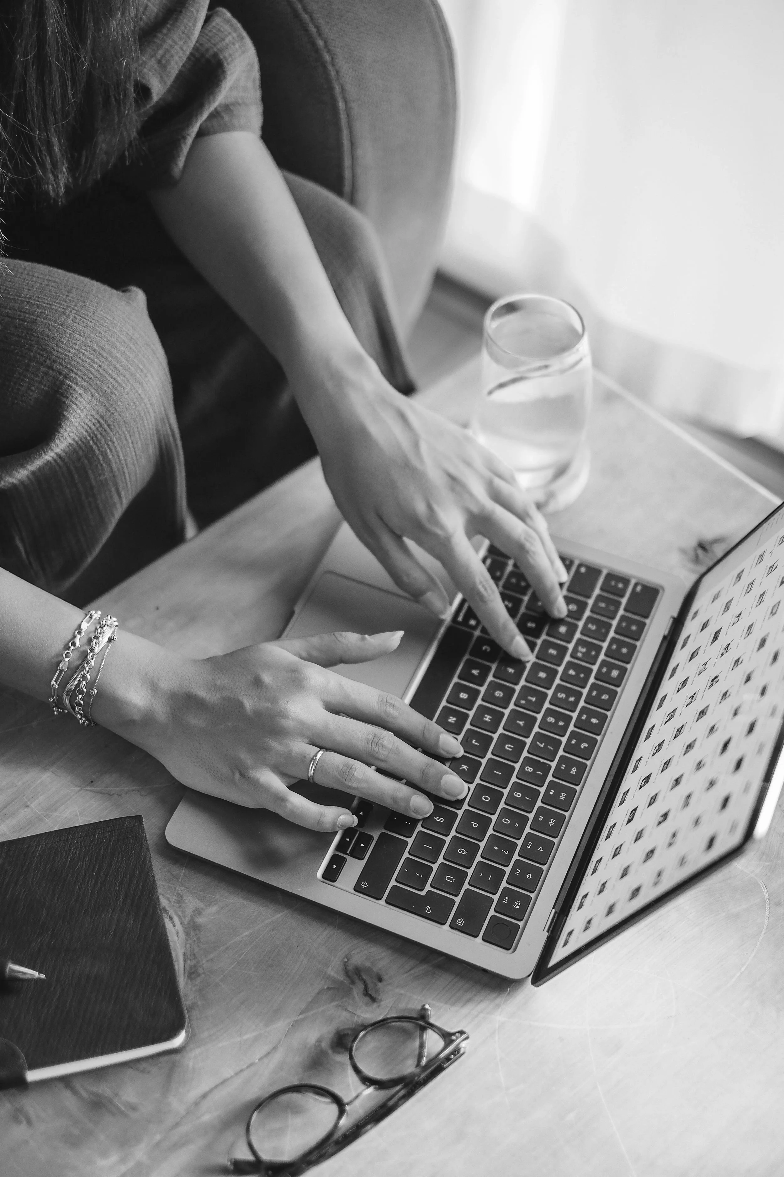 A person typing on a laptop keyboard with a glass of water nearby, glasses, a notebook, and bracelets visible on their wrist.