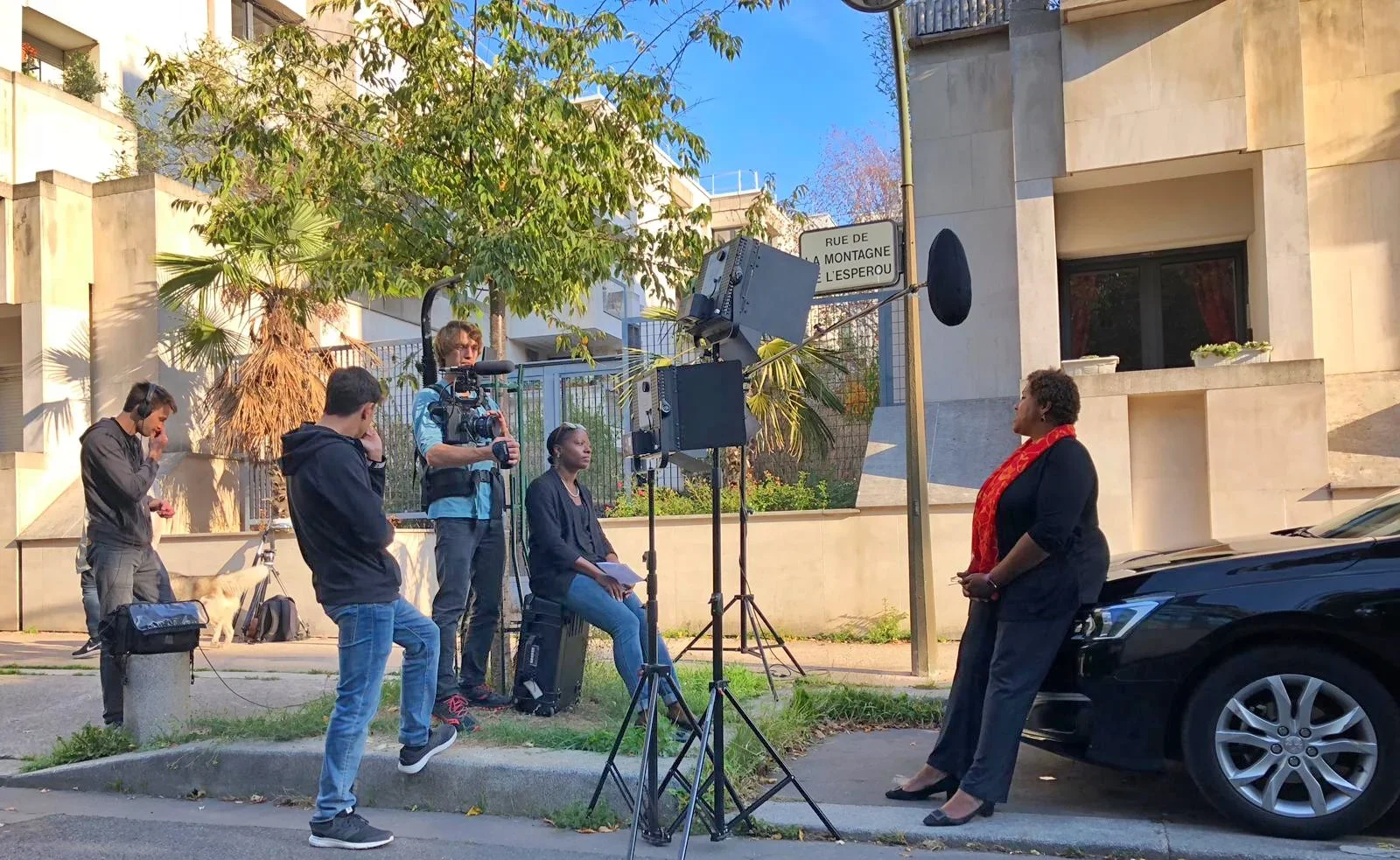 A woman being interviewed or filmed by a camera crew on a sidewalk, with a cameraman operating a professional camera, two crew members, and another person sitting nearby with a dog in the background.