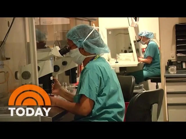 NBC News Today Show: Two scientists or medical professionals in scrubs, wearing protective gear, working in a laboratory with microscopes.
