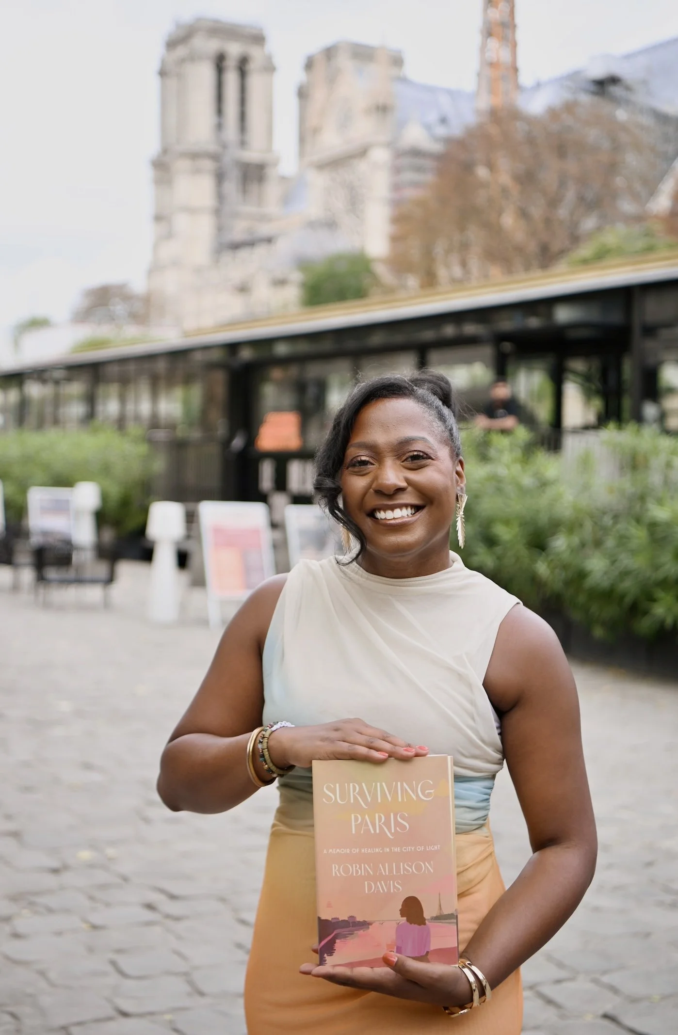A woman smiling and holding a book titled 'Surviving Paris' outdoors in front of a historic building with two towers. She is wearing a light-colored sleeveless top and a yellow skirt.
