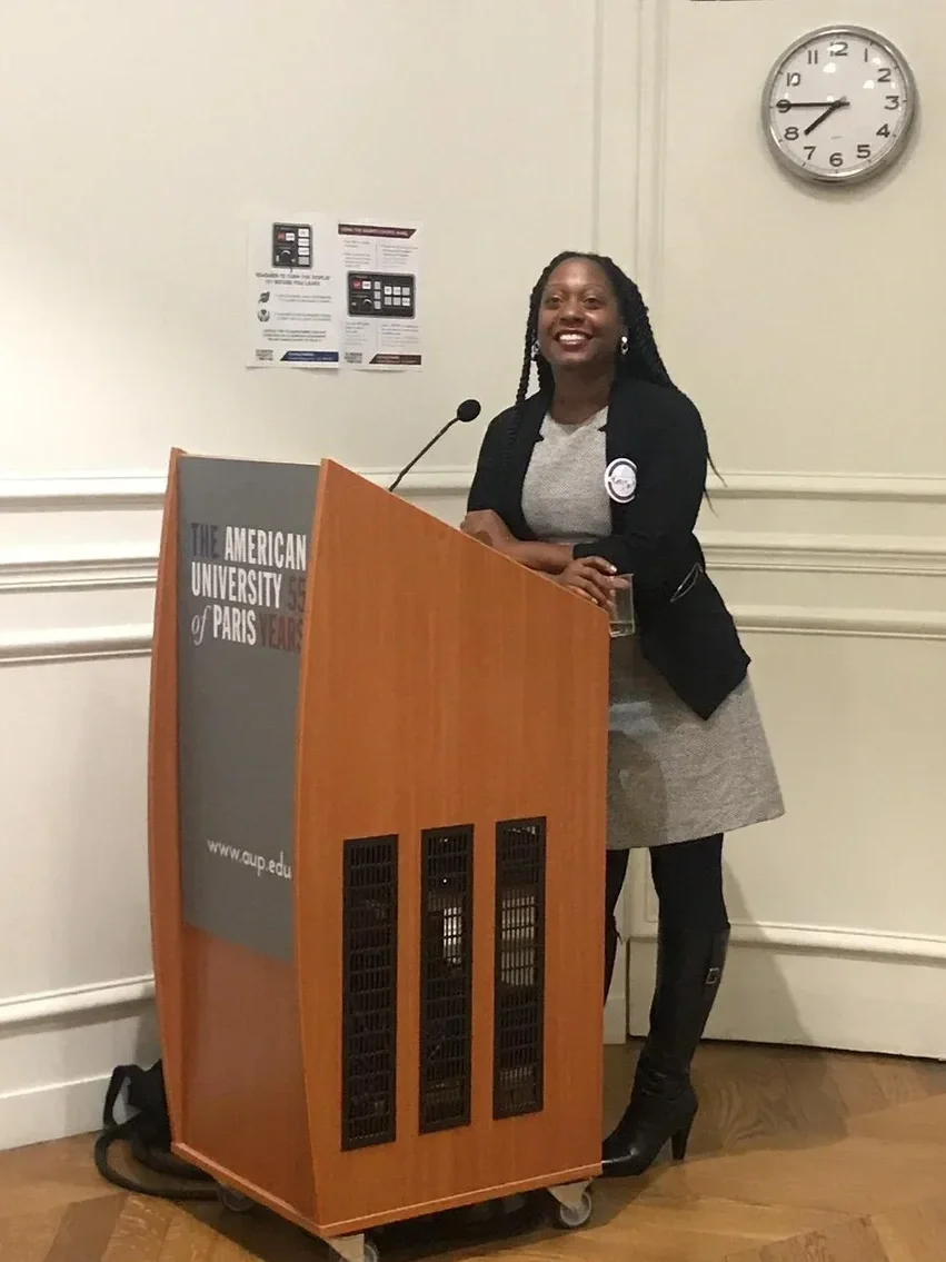  Robin Allison Davis standing behind a wooden podium with a gray front that reads "The American University of Paris". She is smiling, wearing a black blazer, gray dress, and high black boots. 