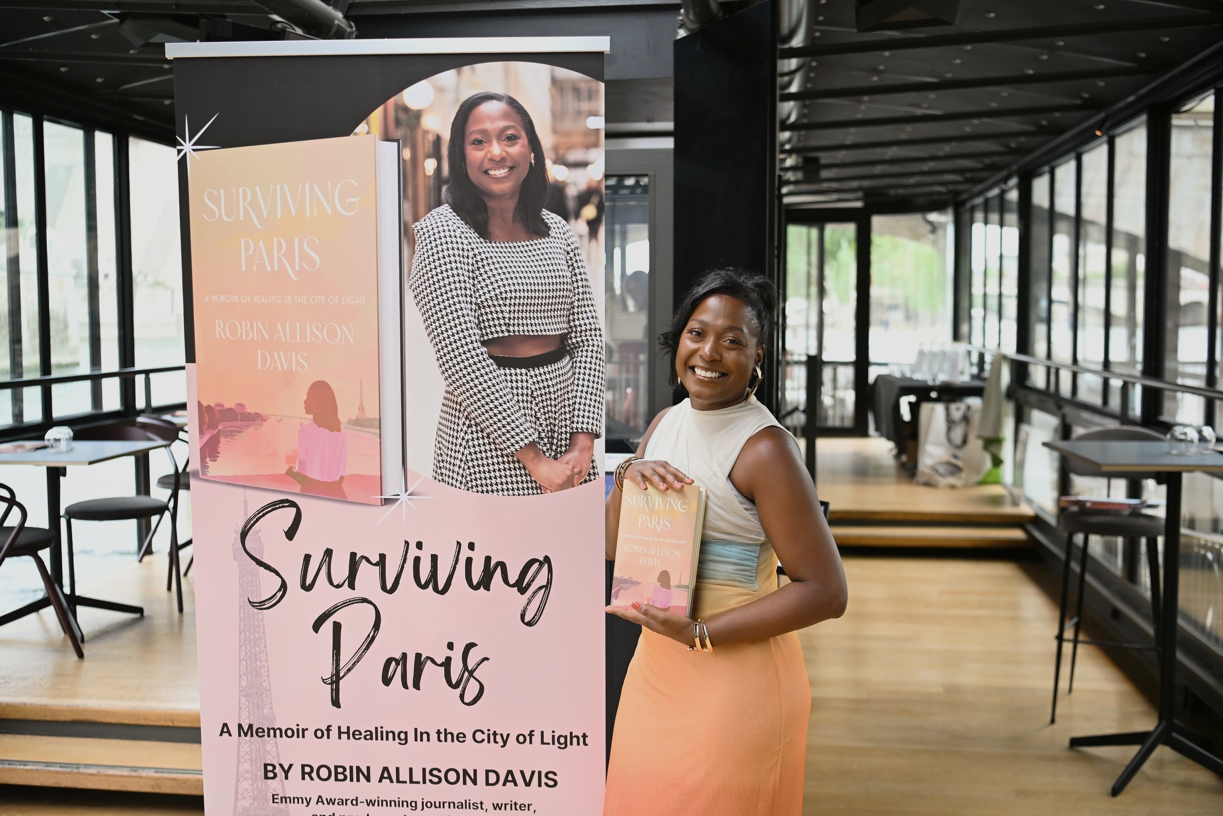 A woman standing next to a promotional poster for the book 'Surviving Paris' by Robin Allison Davis in a cafe or restaurant. The woman is smiling, holding a copy of the book. The poster features a large image of the author and the book cover with a pink and peach background, and a silhouette of the Eiffel Tower.