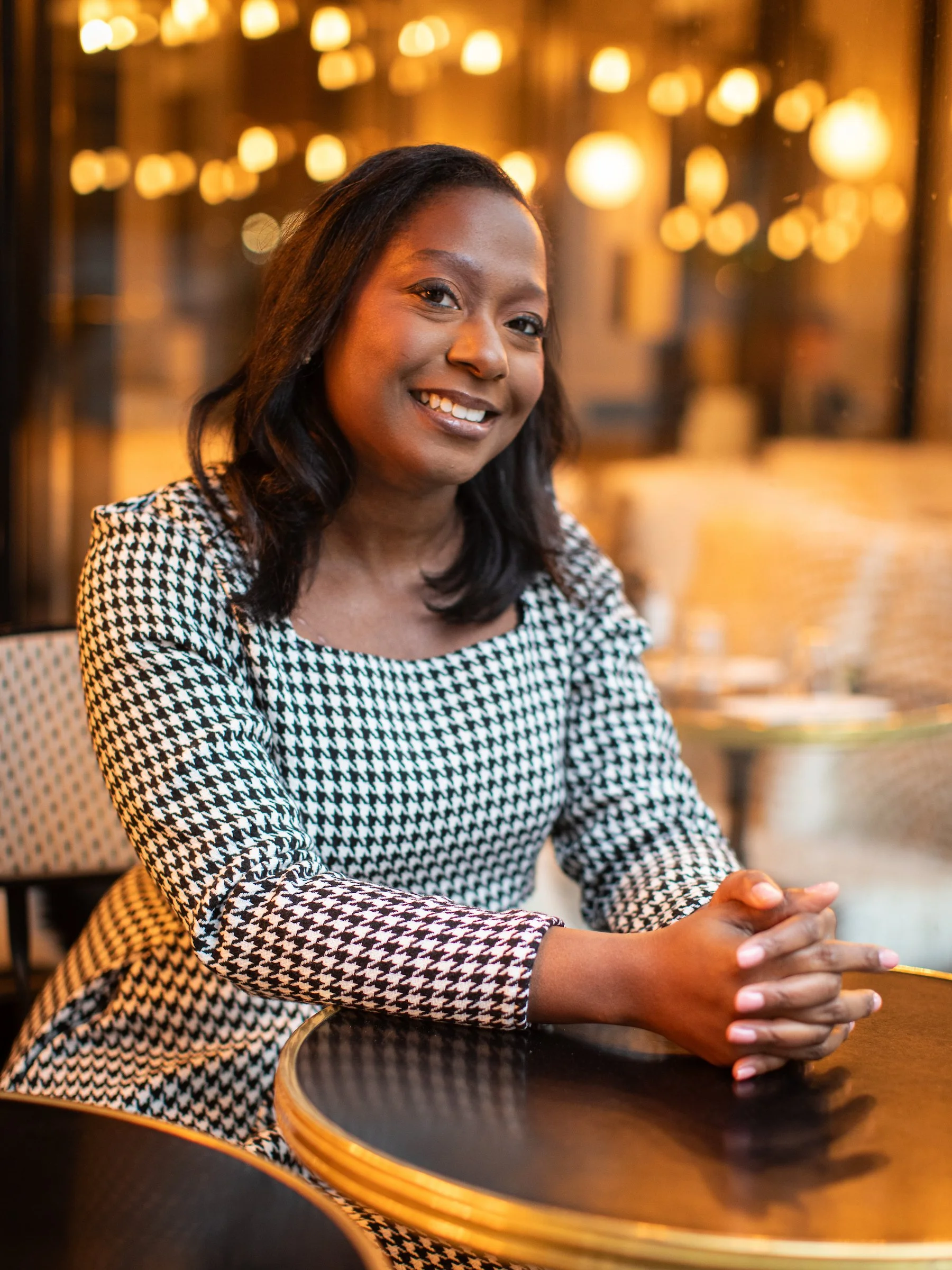 A woman with dark hair wearing a houndstooth patterned dress, sitting at a table in a warmly lit restaurant or cafe, smiling at the camera.